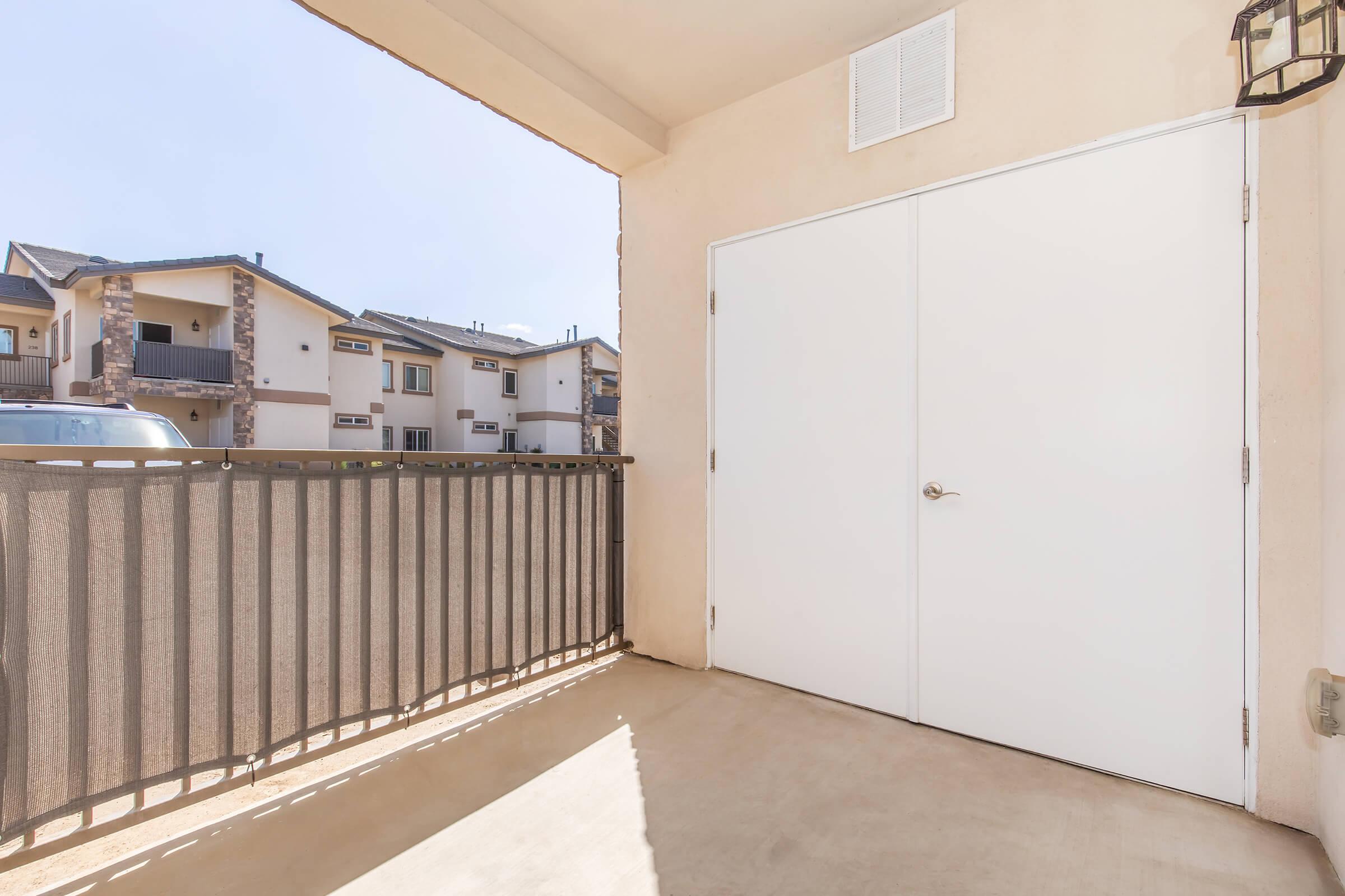 A spacious balcony overlooking a residential complex, featuring a railing and a white door. The exterior wall is painted beige, and there is a clear blue sky above. Sunlight casts shadows on the floor, creating a bright and airy atmosphere.