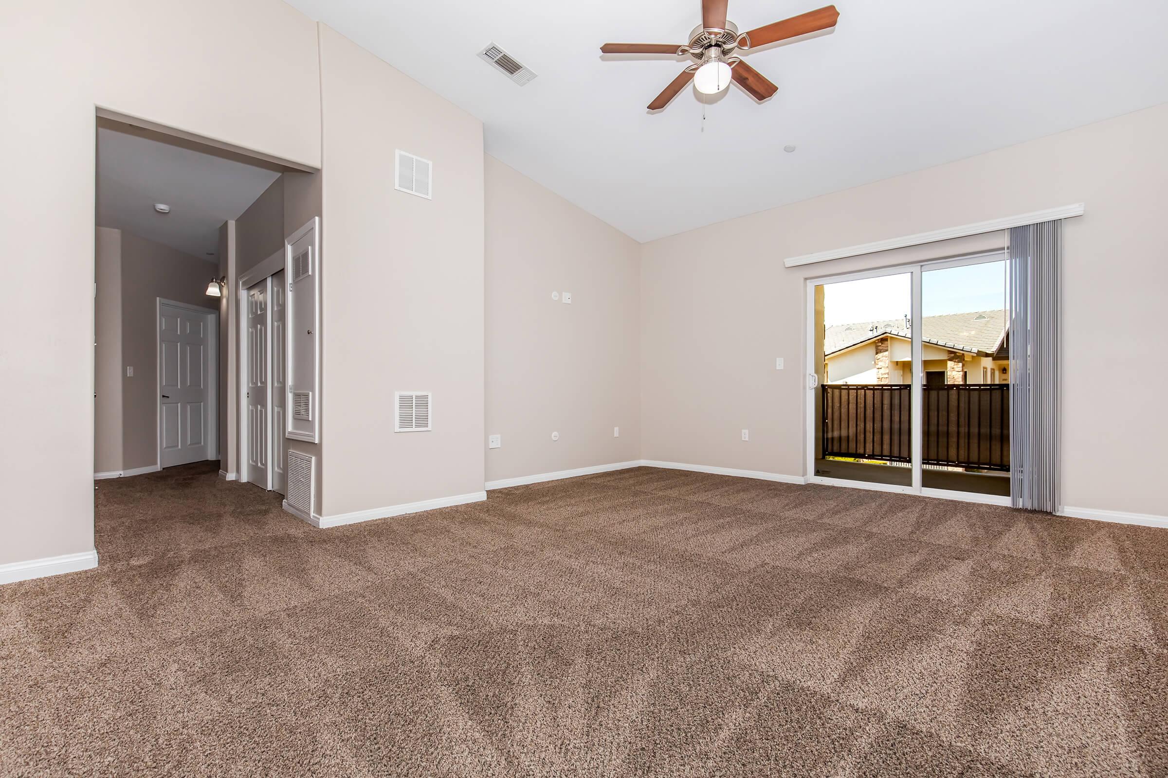 Spacious living room with a ceiling fan, light beige walls, and plush carpet. Large sliding glass door leads to an outdoor balcony. Adjacent hallway and doorways can be seen, contributing to an open layout and bright ambiance. Ideal for relaxing or entertaining.