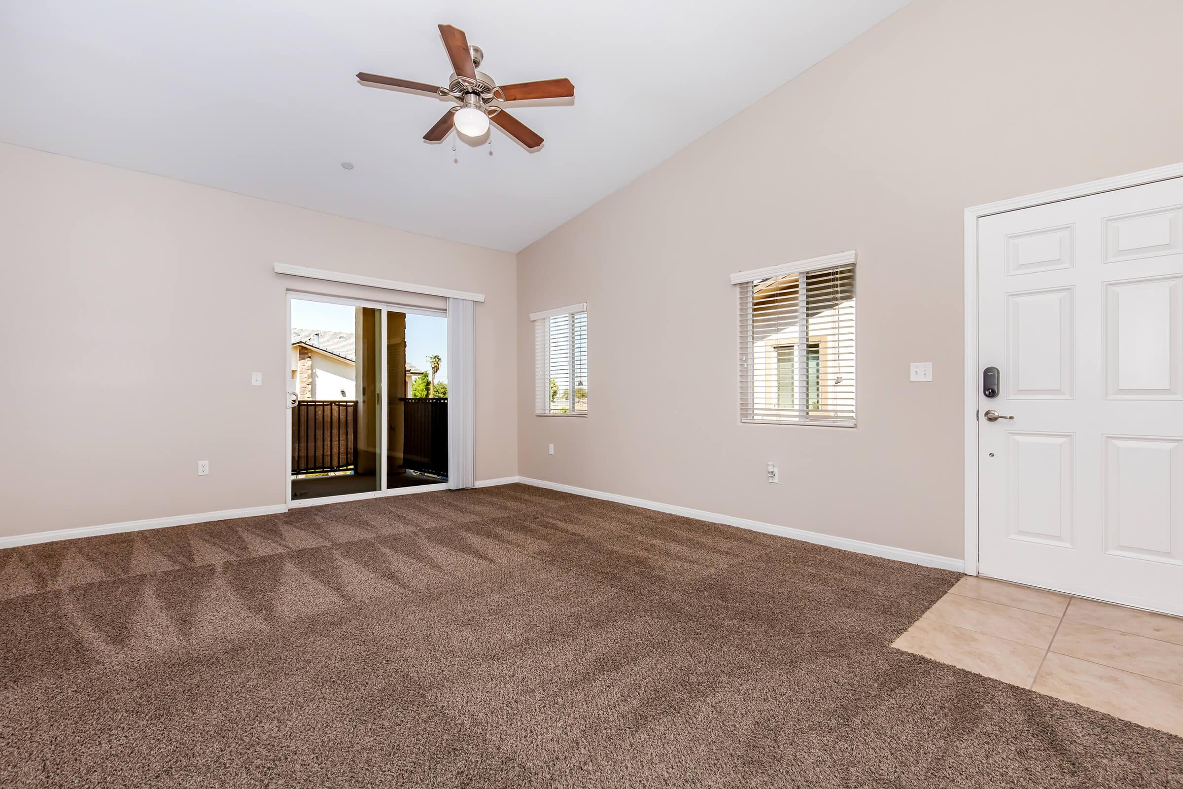 Spacious interior with light-colored walls and a ceiling fan. Features large windows allowing natural light, a sliding glass door leading to a balcony, and a carpeted floor. The room has a front entrance with a white door and tile flooring near the entrance.
