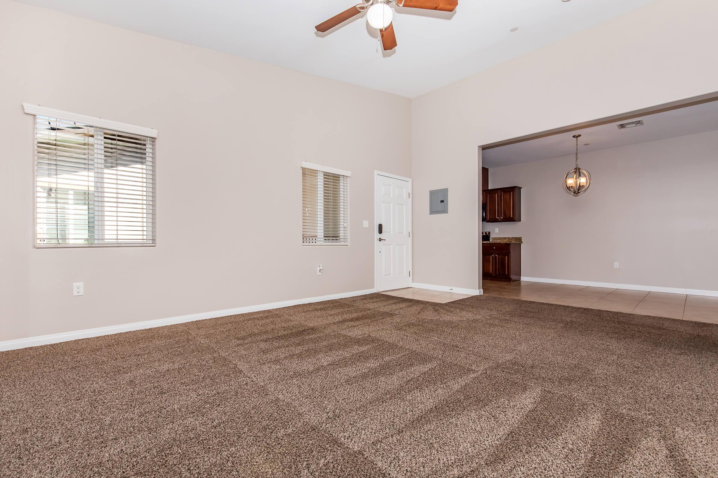 A spacious living area with light-colored walls, a ceiling fan, and large windows featuring blinds. The floor is covered with brown carpet, leading to an open kitchen visible through an archway. The kitchen has wooden cabinets and modern light fixtures, creating a warm, inviting atmosphere.