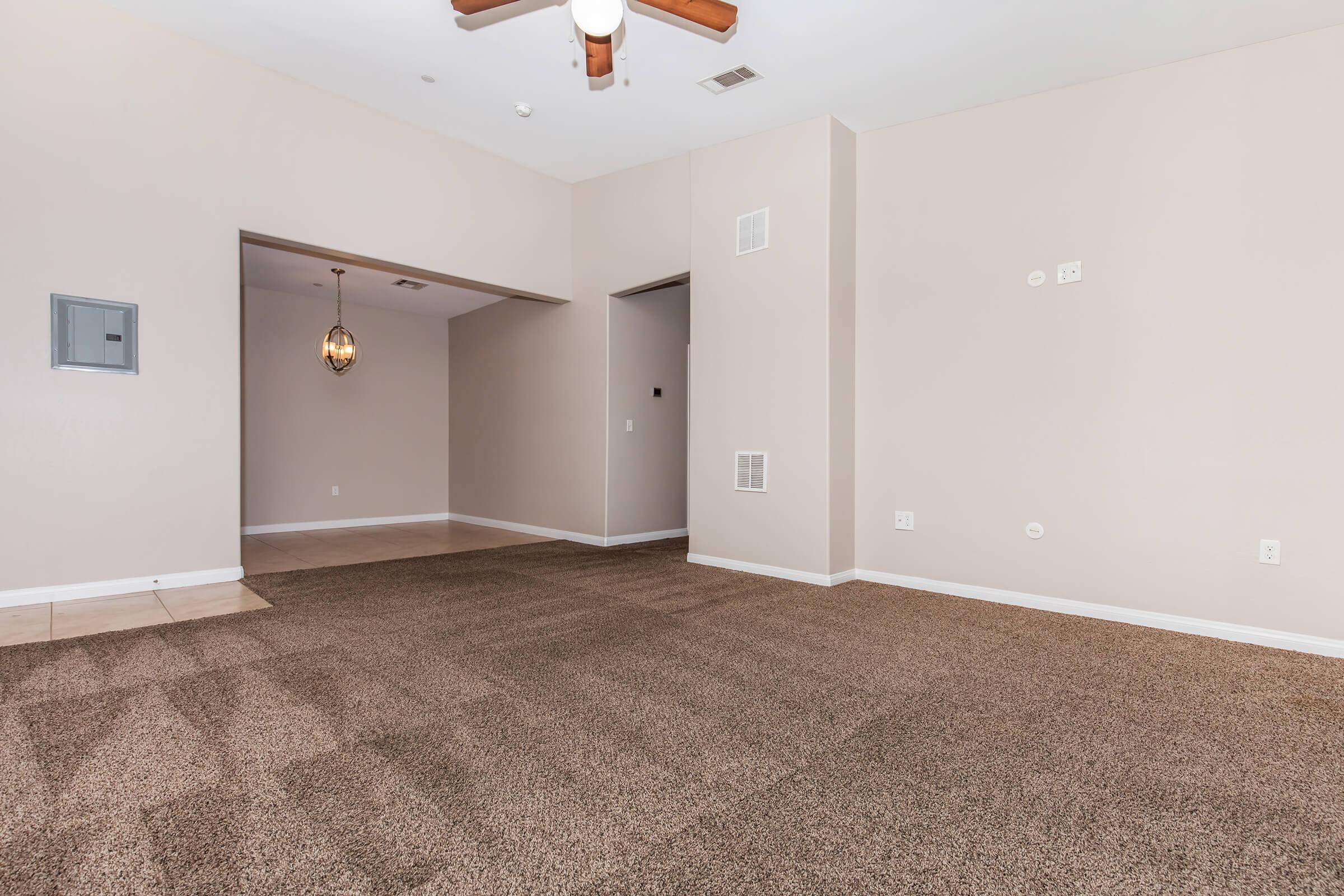 A spacious, empty room with beige walls and a tan carpet. A ceiling fan hangs from the ceiling, and there is a doorway leading to another room. A chandelier is visible in the adjoining space, providing a warm ambiance. The area is well-lit, creating an inviting atmosphere.