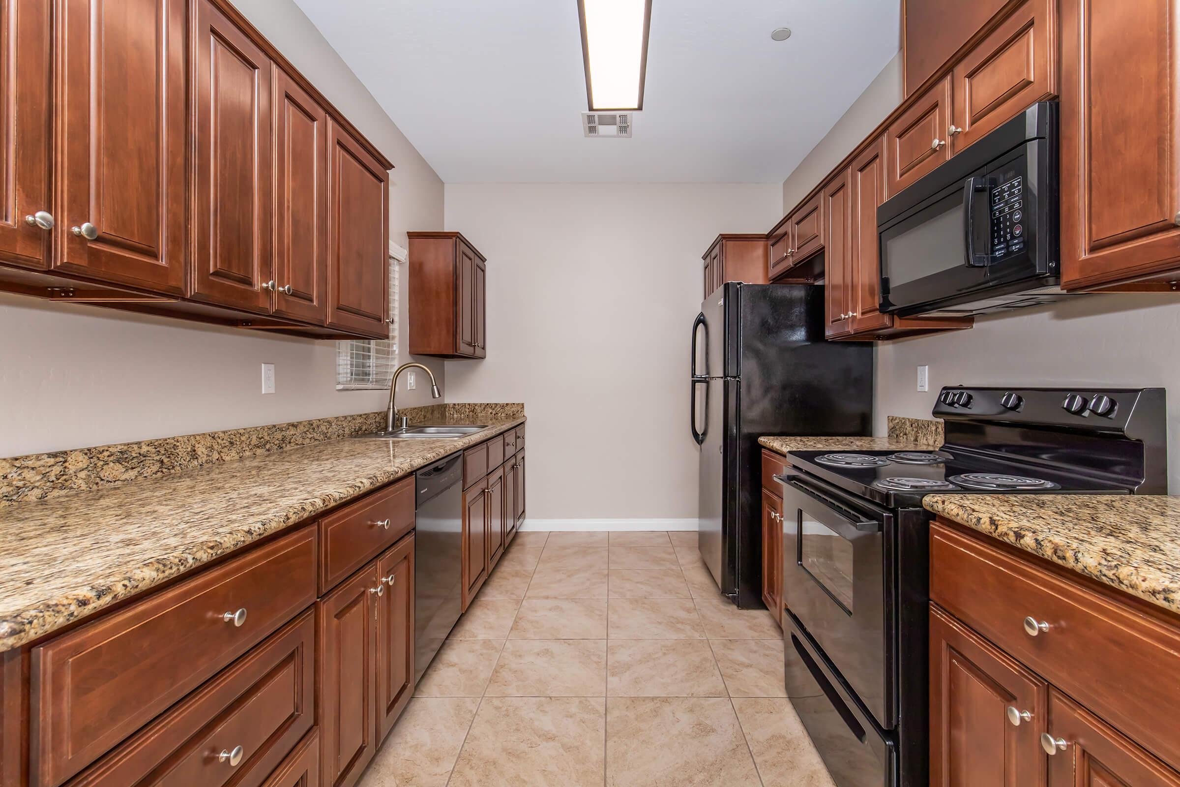 A modern kitchen featuring dark wooden cabinets, granite countertops, and stainless steel appliances. The space includes a stove, oven, microwave, refrigerator, and a sink. The floor is tiled, and the walls are a neutral color, creating a clean and spacious appearance.
