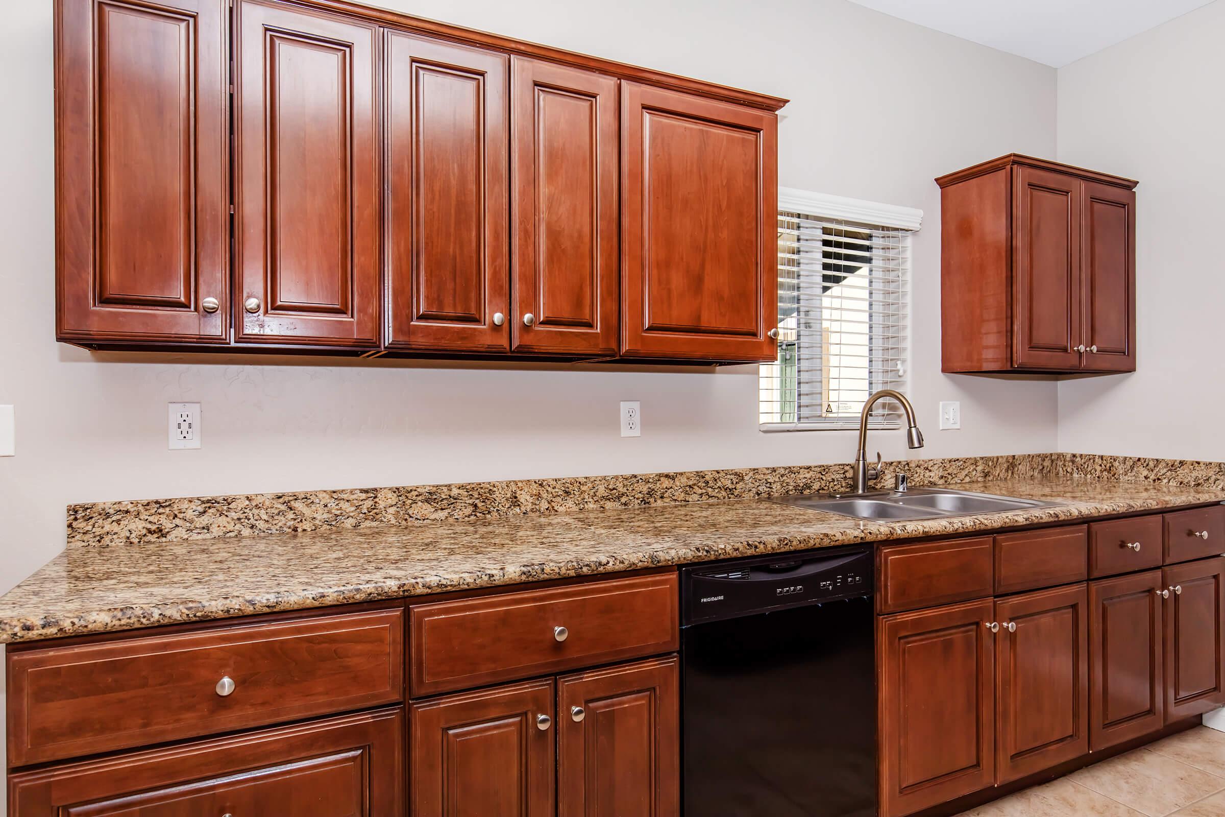 Modern kitchen featuring wooden cabinets in a rich brown finish, a granite countertop, and a double sink with a single handle faucet. A black dishwasher is installed beneath the countertop, and there is a window with blinds that lets in natural light. The walls are painted in a light color, enhancing the space.