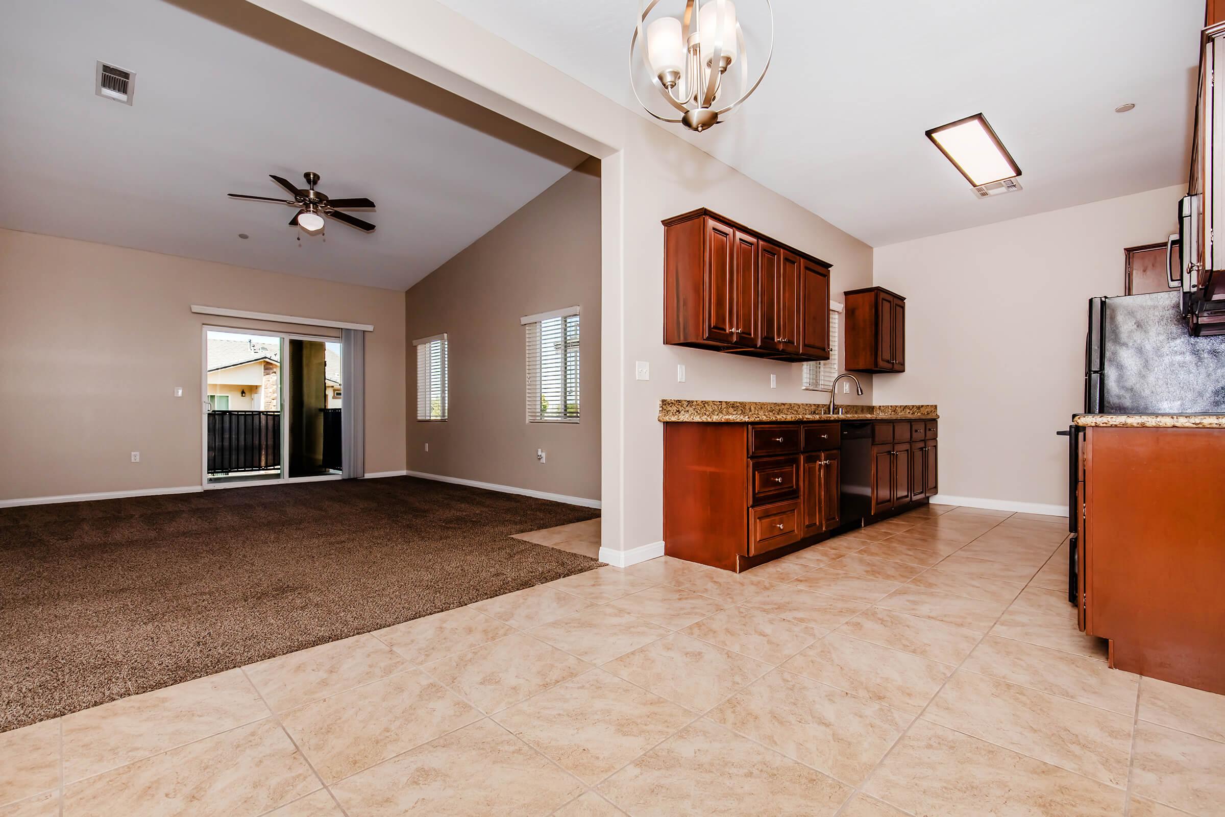 Interior view of a modern home featuring a spacious kitchen with dark wooden cabinets and granite countertops. The living area has a high ceiling, a ceiling fan, and is carpeted, with windows allowing natural light. The layout shows an open design connecting the kitchen and living spaces.