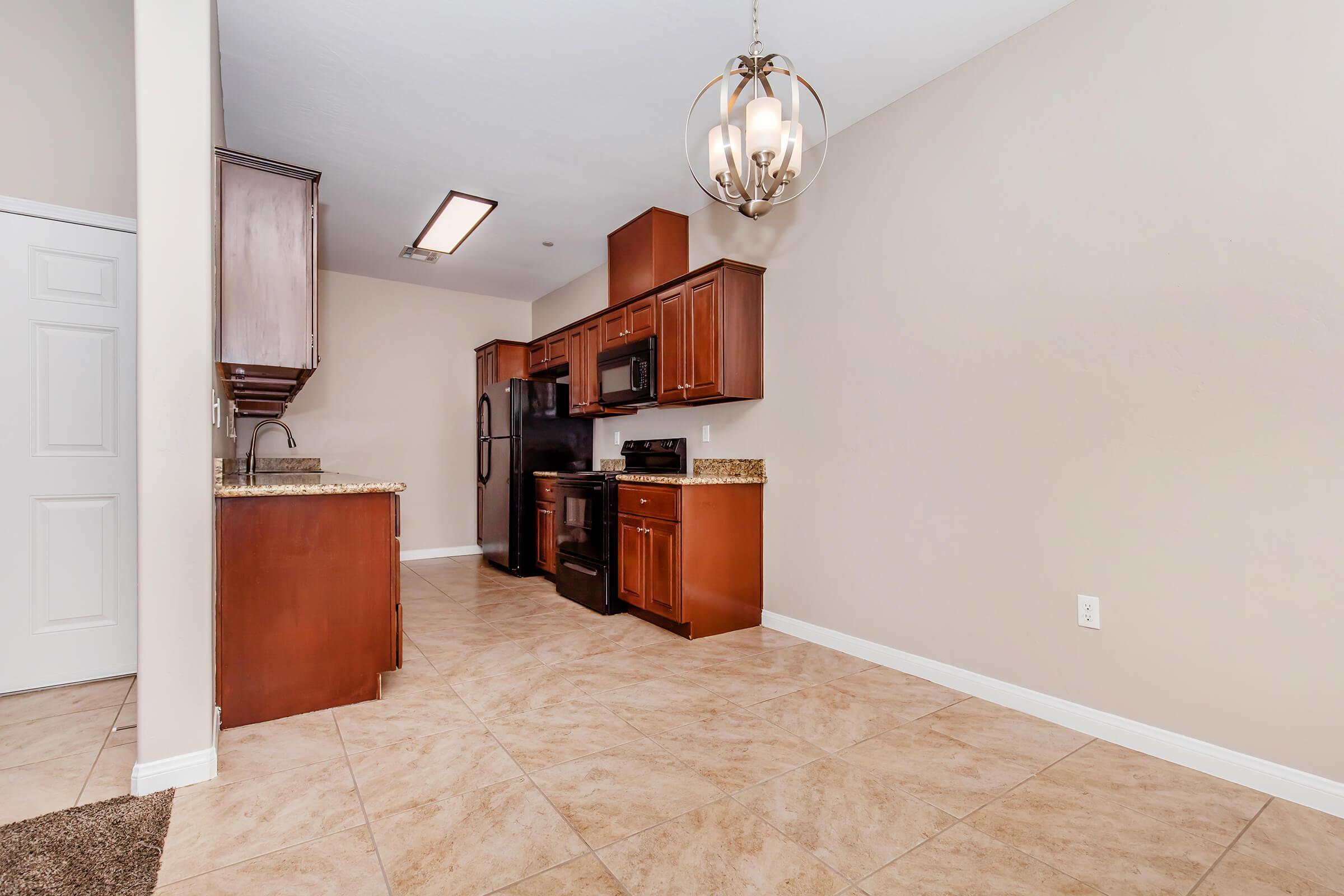 A modern kitchen featuring dark wood cabinetry, black appliances, and granite countertops. The space has a neutral wall color and tile flooring, with a stylish pendant light fixture hanging above the kitchen island. A doorway is visible in the background.