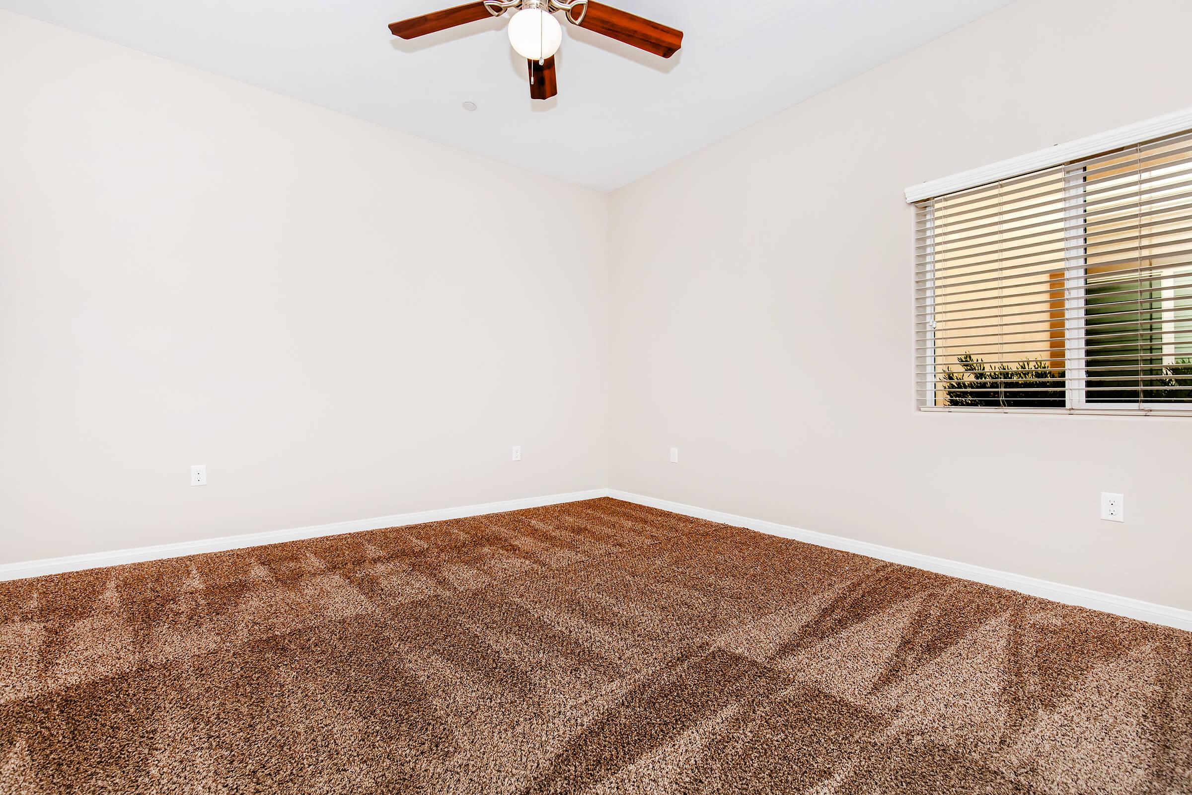 Empty room with beige walls and a light carpet. A ceiling fan with wooden blades is visible. There is a window with horizontal blinds allowing natural light to enter, casting shadows on the carpeted floor. The overall ambiance is bright and uncluttered, suggesting a fresh living space.