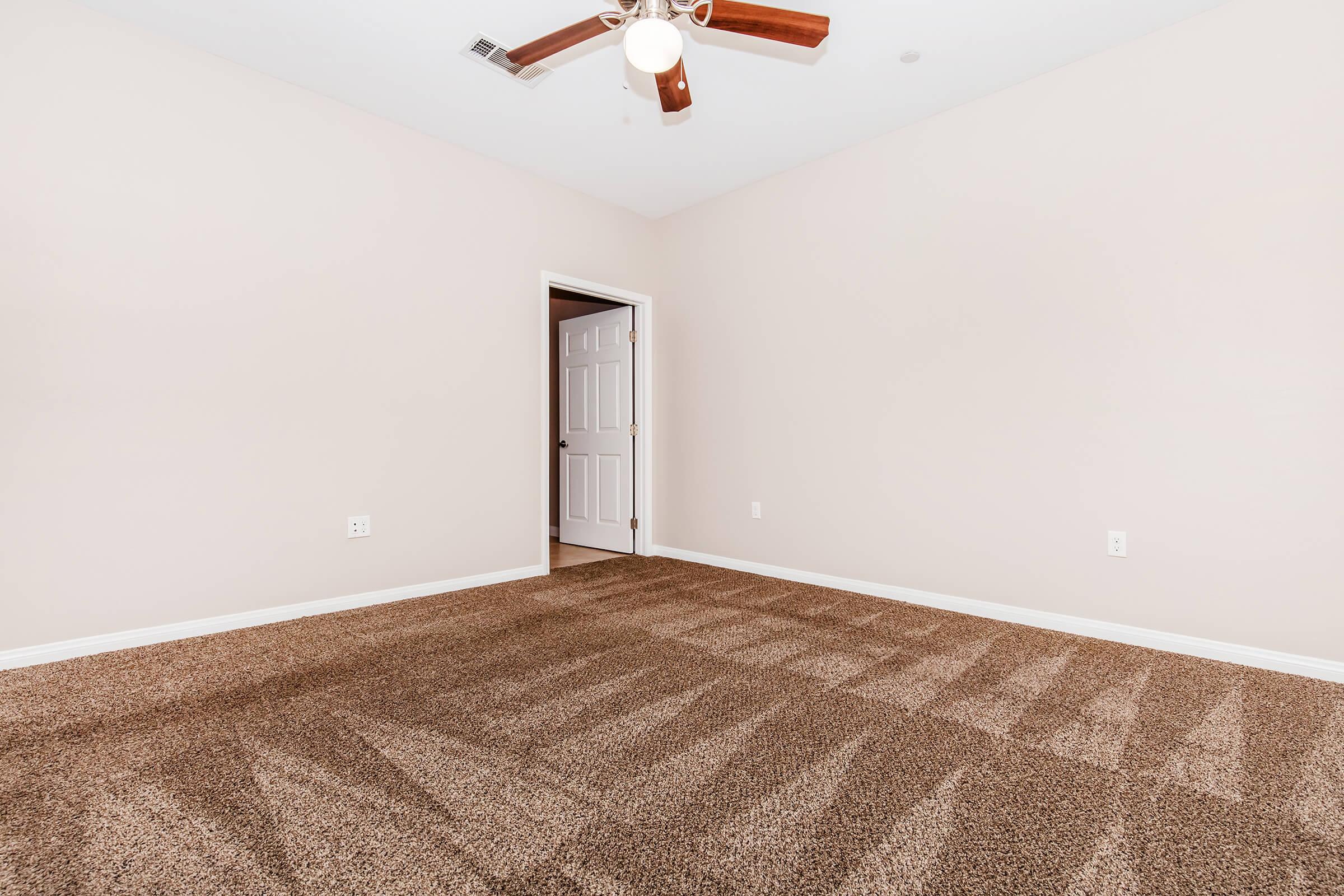 A spacious, empty room with light beige walls and a ceiling fan. The floor is covered in plush brown carpet, and there is a partially open white door leading to another area. The room is well-lit, creating an inviting atmosphere.