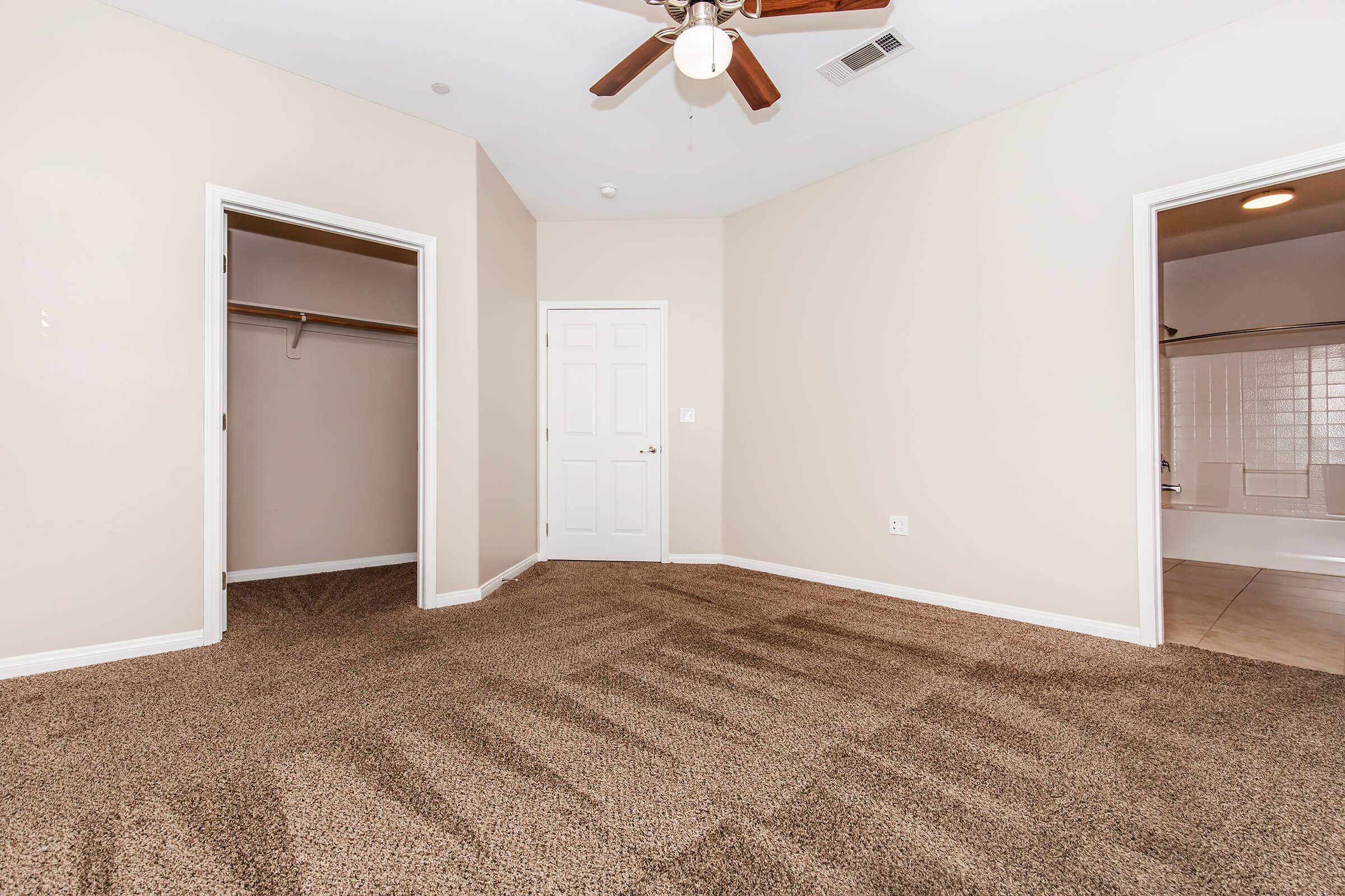 A spacious, empty room with beige walls and carpet flooring. It features a ceiling fan, a closet on the left, and a doorway leading to a bathroom on the right. Natural light enters from the open door, creating a bright atmosphere.