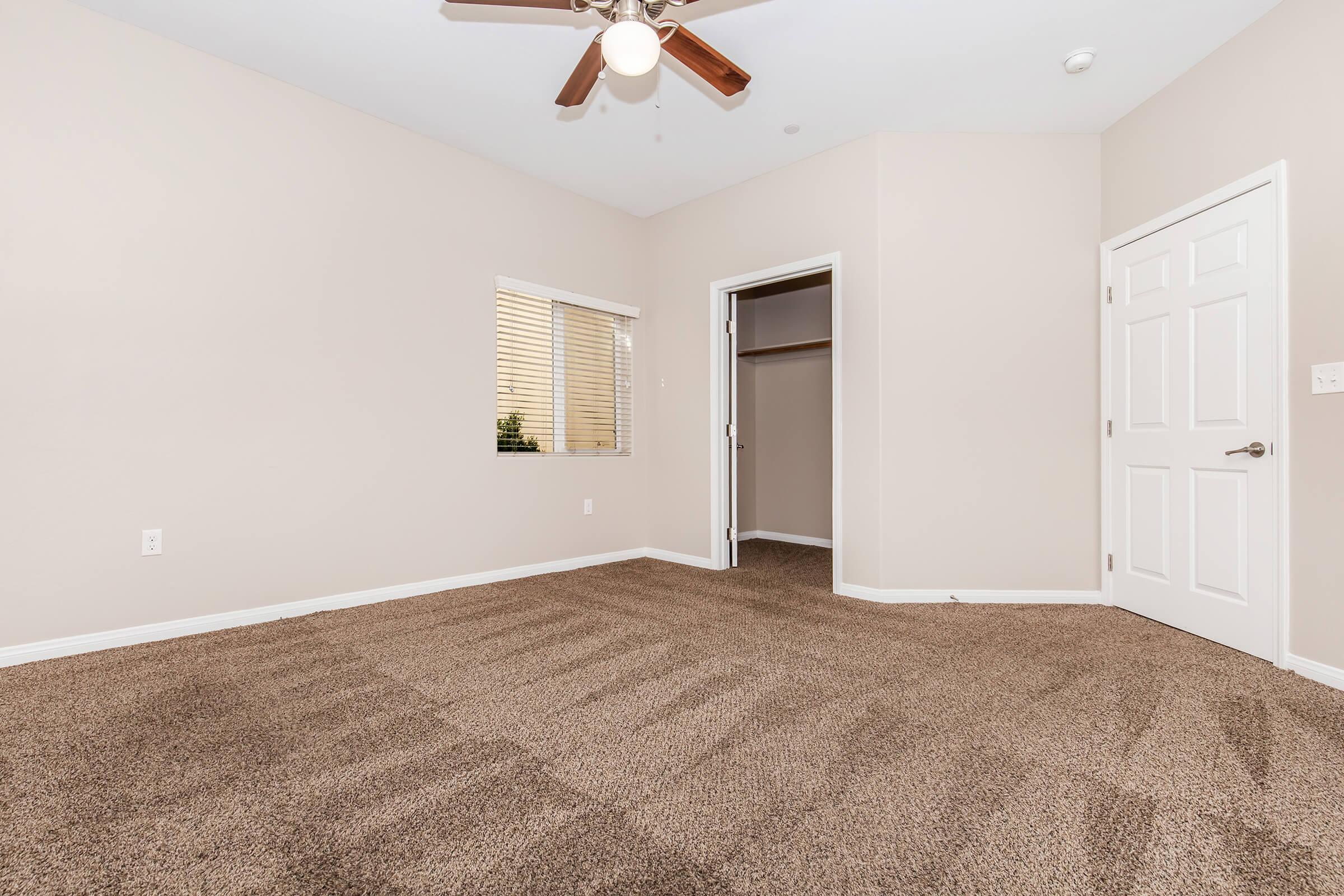 A spacious room with light beige walls and a ceiling fan. The floor is covered in brown carpet with visible patterns. There is a window allowing natural light in, and a closet door visible on the right. The room is empty, creating a bright and airy atmosphere.