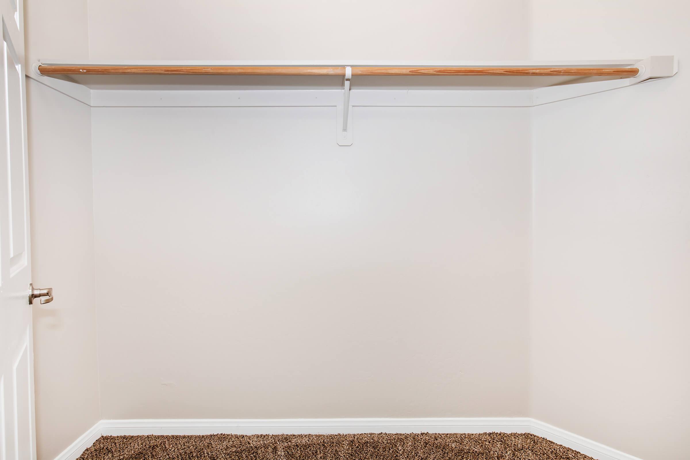 A clean, empty closet featuring a wooden hanging rod mounted on a white wall. The floor is covered with brown carpet, and there is a white door on the left side of the image. The overall appearance is minimal and uncluttered.