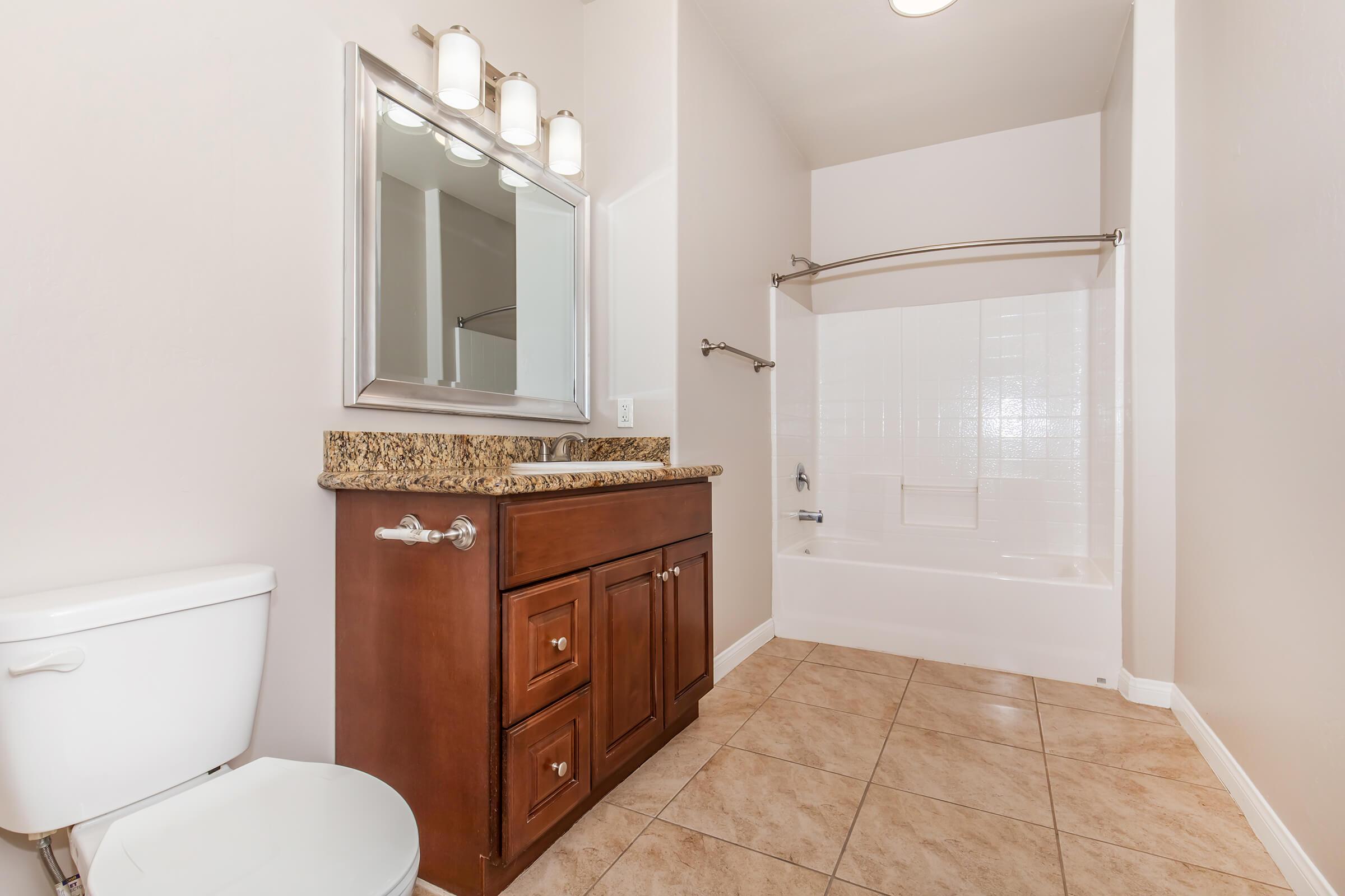 A modern bathroom featuring a wooden vanity with a granite countertop and a rectangular mirror above it. There is a toilet on the left and a shower/tub combo with a glass shower rod on the right. The walls are painted in a light color, and the floor is tiled with neutral-colored tiles.