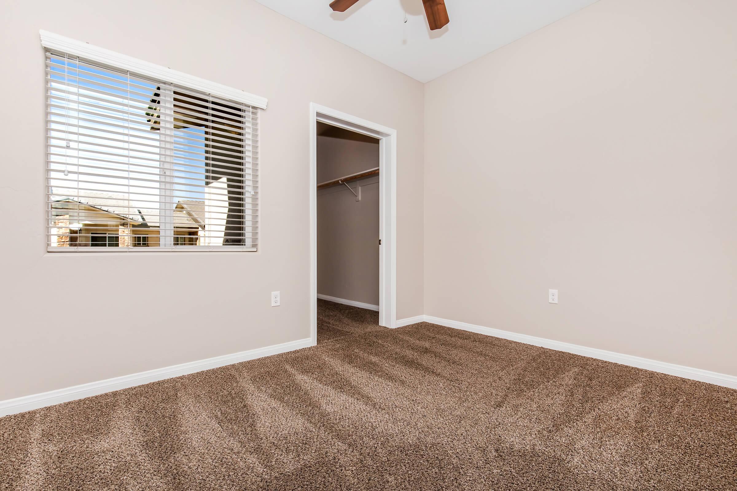 Empty room with light brown carpet and beige walls. A window with white blinds allows natural light to enter, creating a bright atmosphere. A closet is visible through a door on the right side. The room has a ceiling fan, adding to the spacious and airy feel.