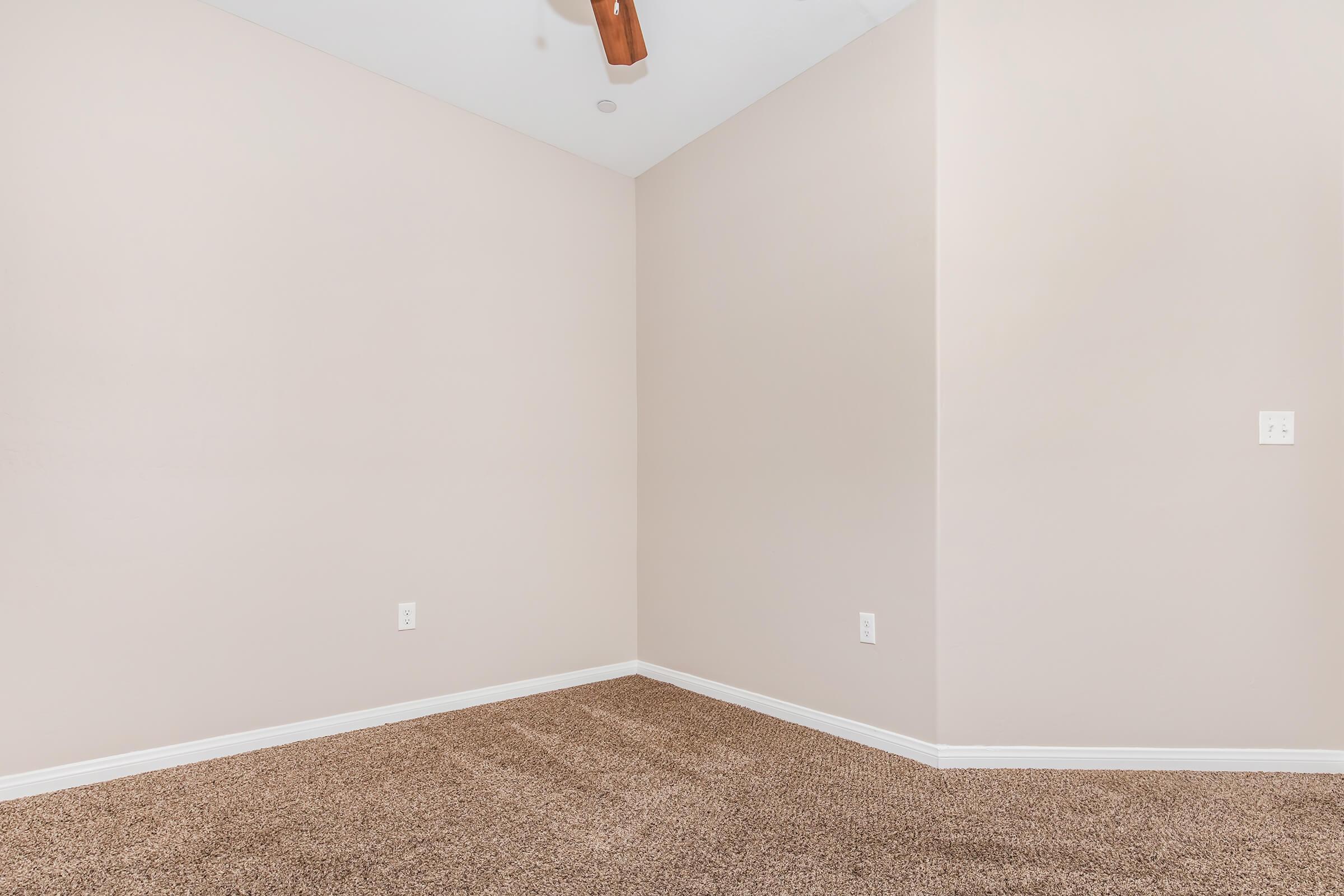 Empty interior corner of a room with light beige walls and a tan carpeted floor. The space has a ceiling fan and no furniture or decorations, creating a minimalistic appearance. The room is well-lit and appears freshly painted.