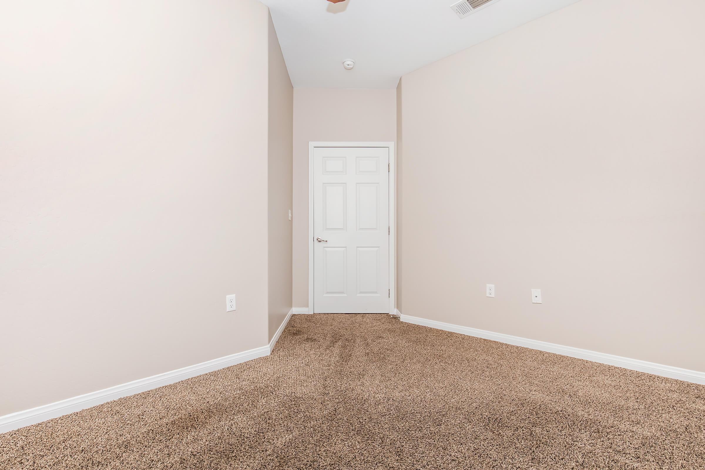 Empty room with beige walls and light brown carpet. A white door is visible on one wall, and there are no furnishings or decorations. The room has a quiet, minimalistic appearance, suitable for various uses.