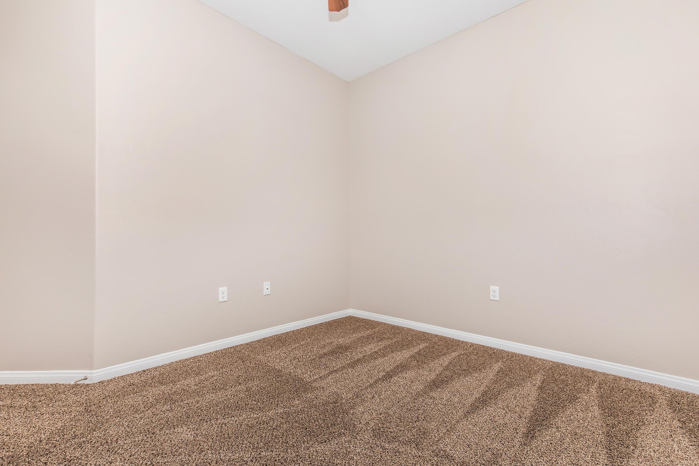 Empty interior corner of a room with light beige walls and carpeted flooring. Two electrical outlets are visible on the wall. A ceiling fan is not shown but implied by the ceiling height. Natural light casts soft shadows on the floor. The space appears clean and ready for furnishing.