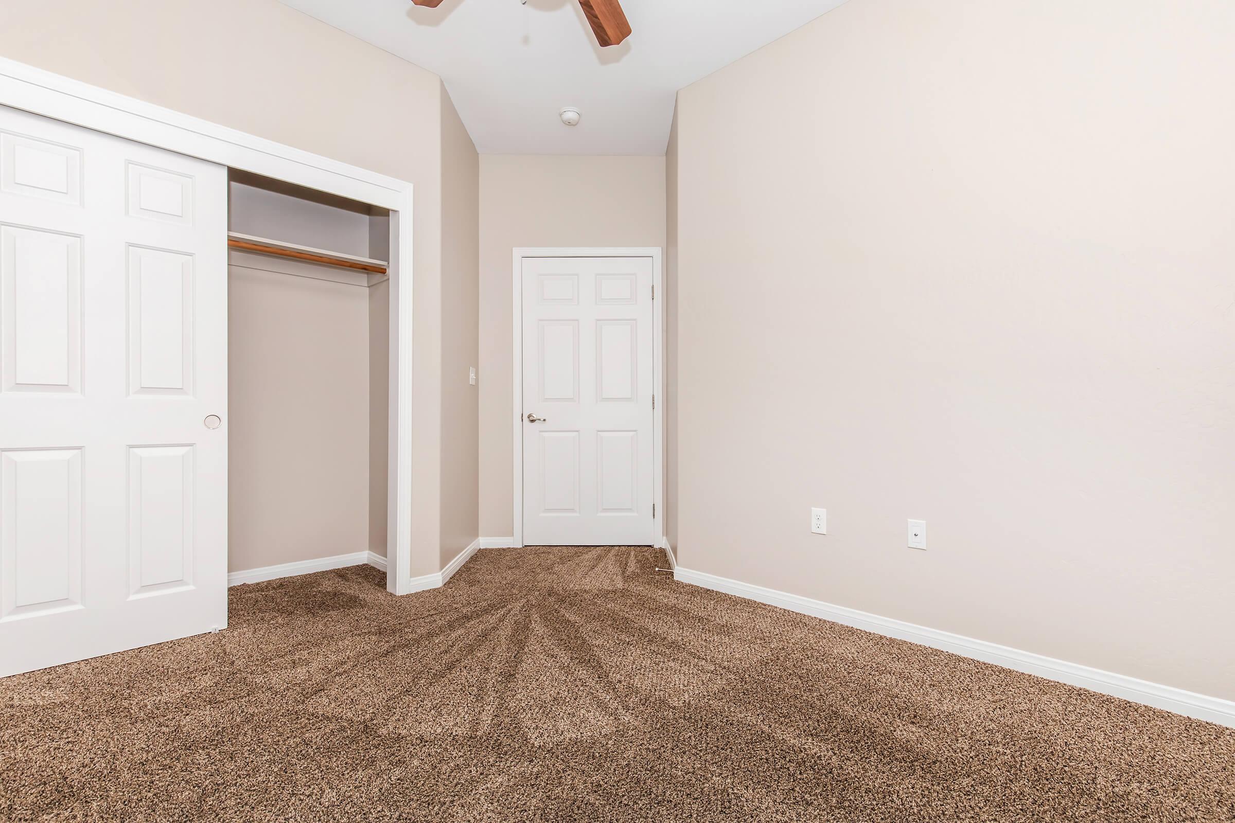 A light-colored room featuring beige walls and plush brown carpeting. There are two doors: one leading to a closet and another to a hallway. The ceiling has a decorative fan, and the space appears well-lit and tidy, suitable for use as a bedroom or office.