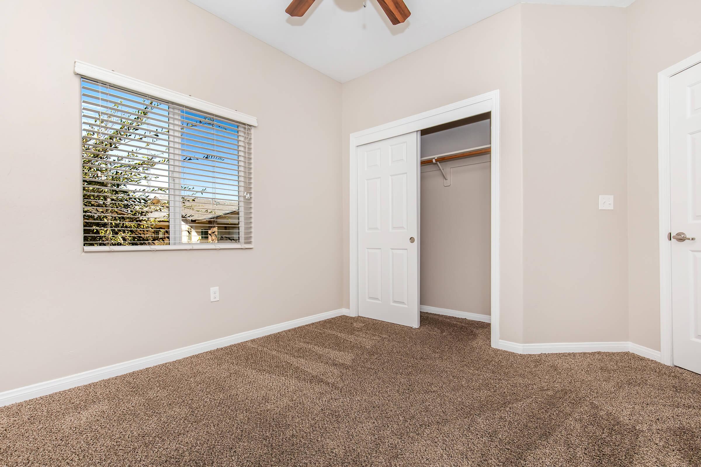 A well-lit, empty room featuring beige walls, a ceiling fan, and a carpeted floor. There is a window with blinds allowing natural light in, and a closet with double doors. The room appears spacious and clean, ideal for various uses such as a bedroom or office.