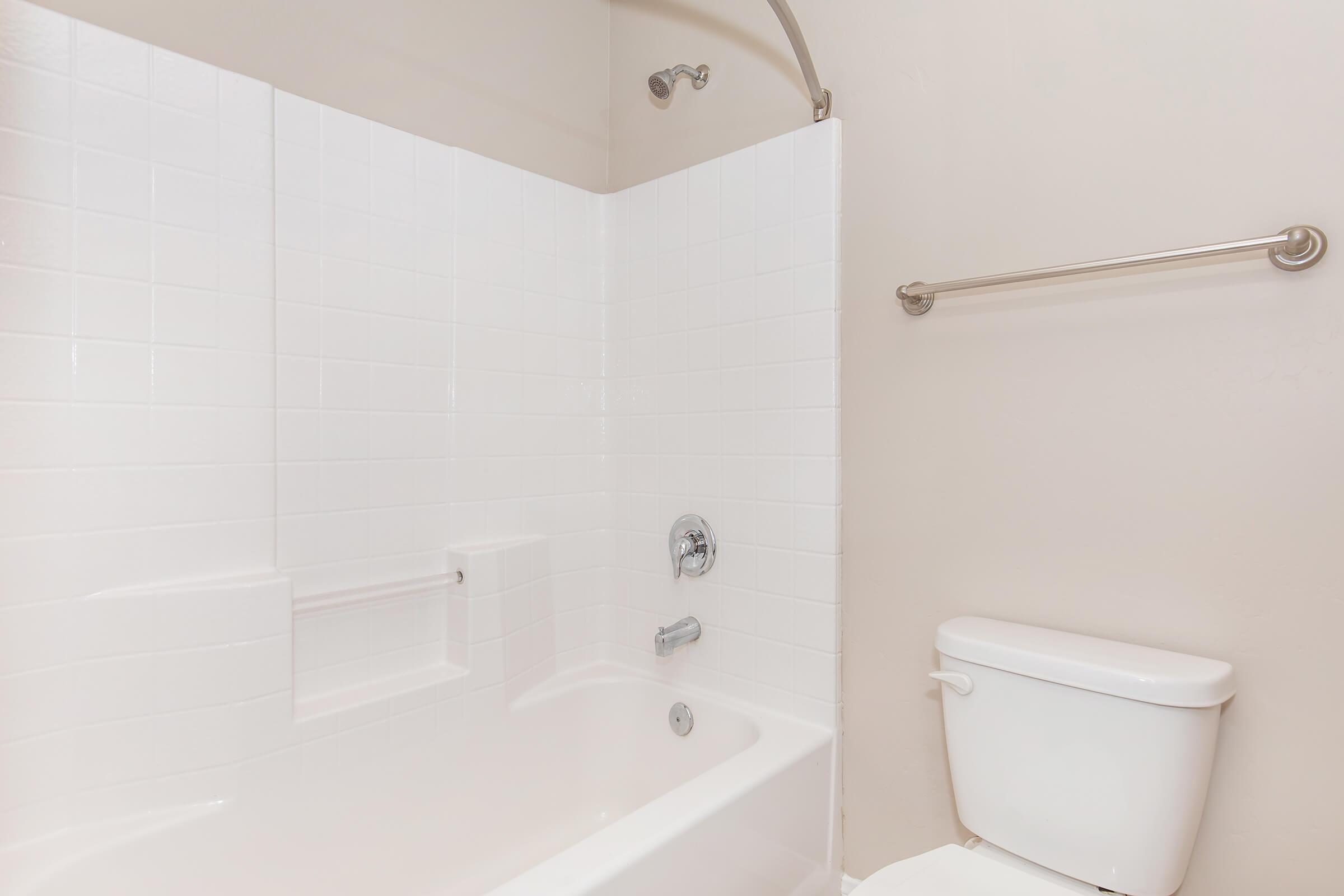 A clean bathroom featuring a white bathtub with a showerhead mounted above, surrounded by white tiled walls. There is a chrome towel bar on the wall and a white toilet beside the bathtub. The overall aesthetic is bright and modern, with a neutral-colored wall.