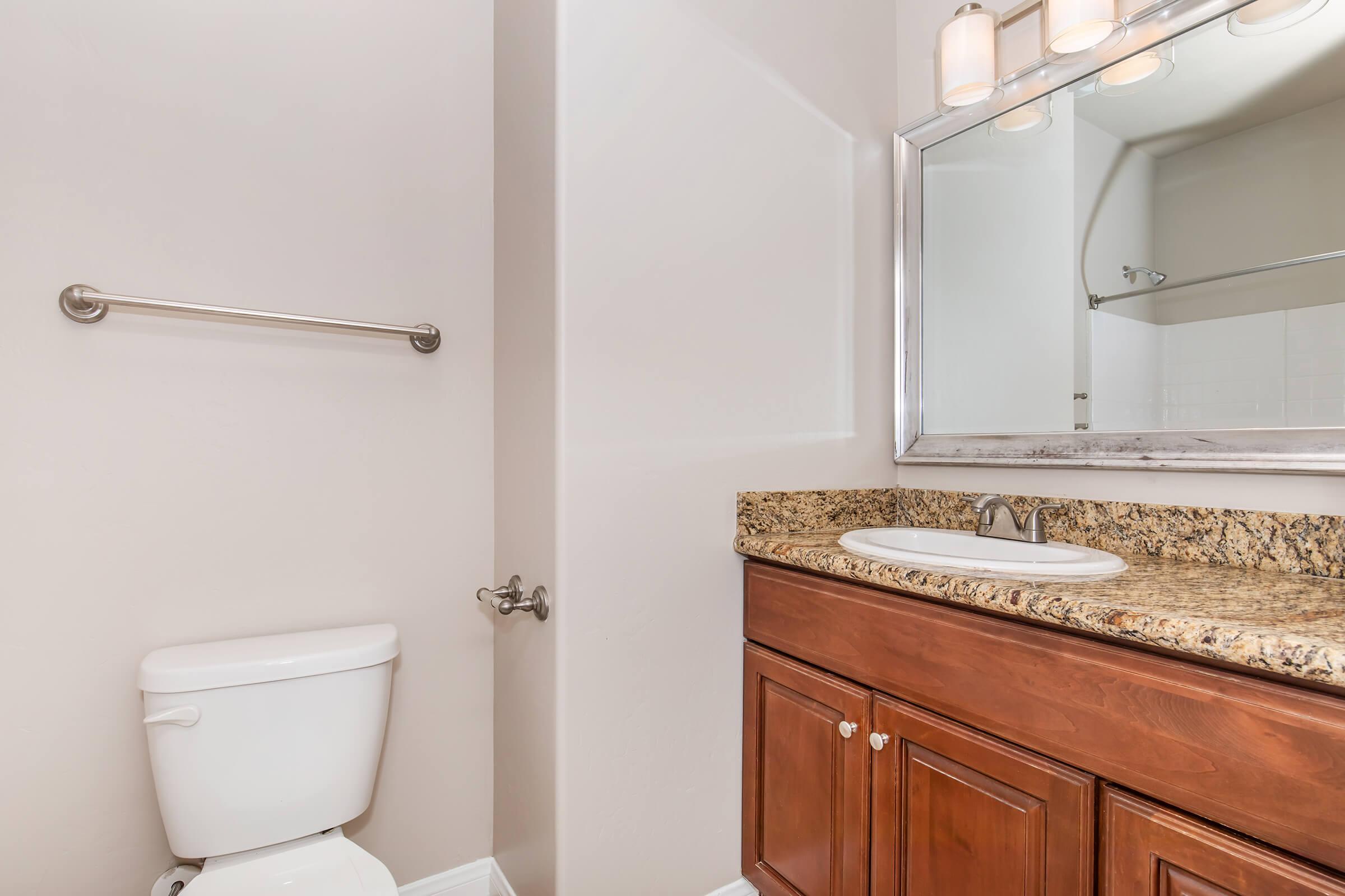 A modern bathroom featuring a white toilet, a wooden vanity with a granite countertop, and a large mirror above the sink. The walls are painted in a light color, and there is a towel rack on one side. The space appears clean and well-lit, contributing to a stylish and functional design.