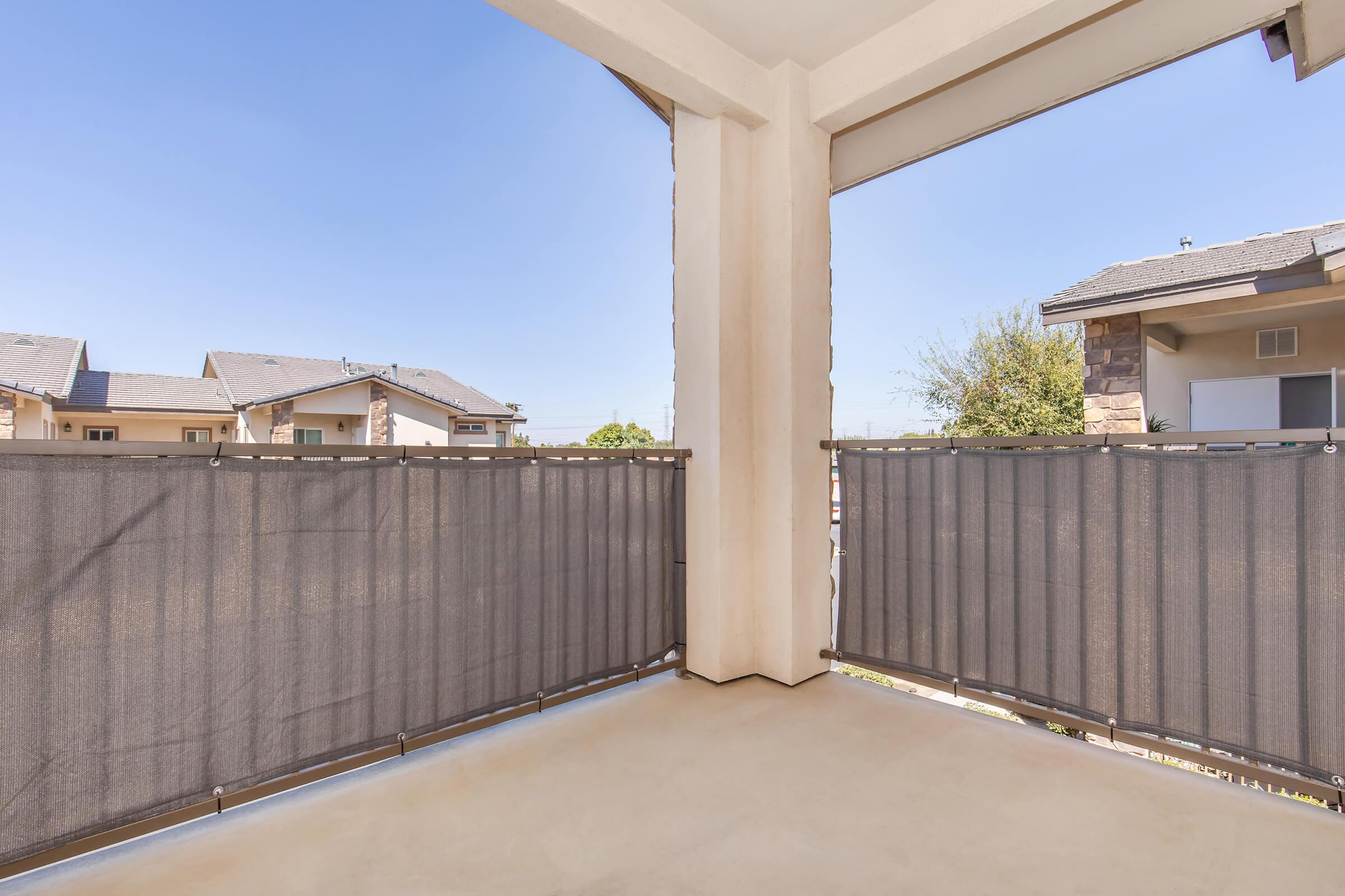A spacious balcony featuring beige flooring and gray privacy screens on either side. In the background, residential buildings and a clear blue sky are visible, providing a bright and open atmosphere.