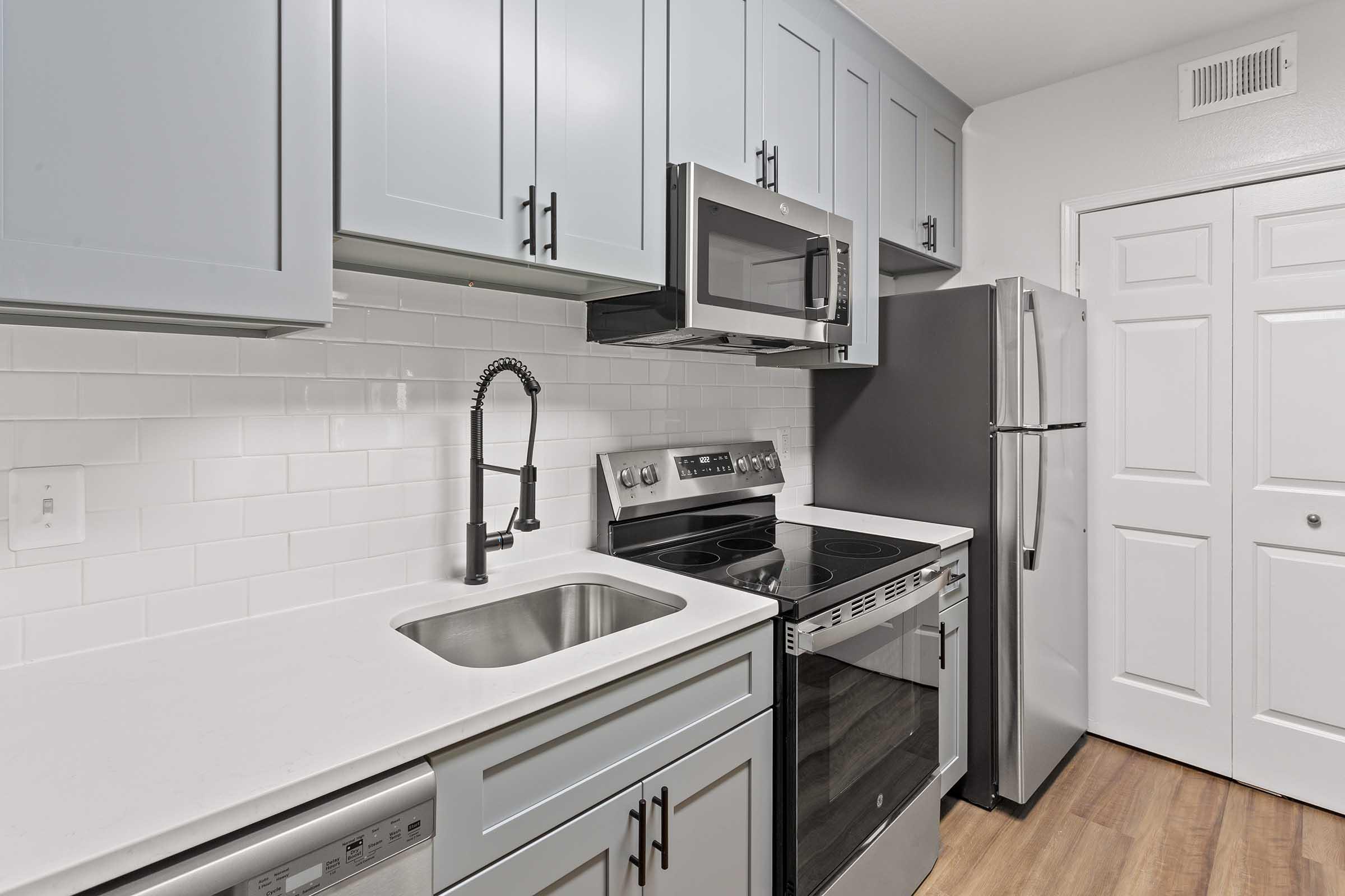 Modern kitchen featuring light blue cabinets, stainless steel appliances, including an oven, microwave, and refrigerator. A single basin sink with a stylish black faucet is situated on a white countertop. The kitchen has white subway tile backsplash and a door leading to another room.