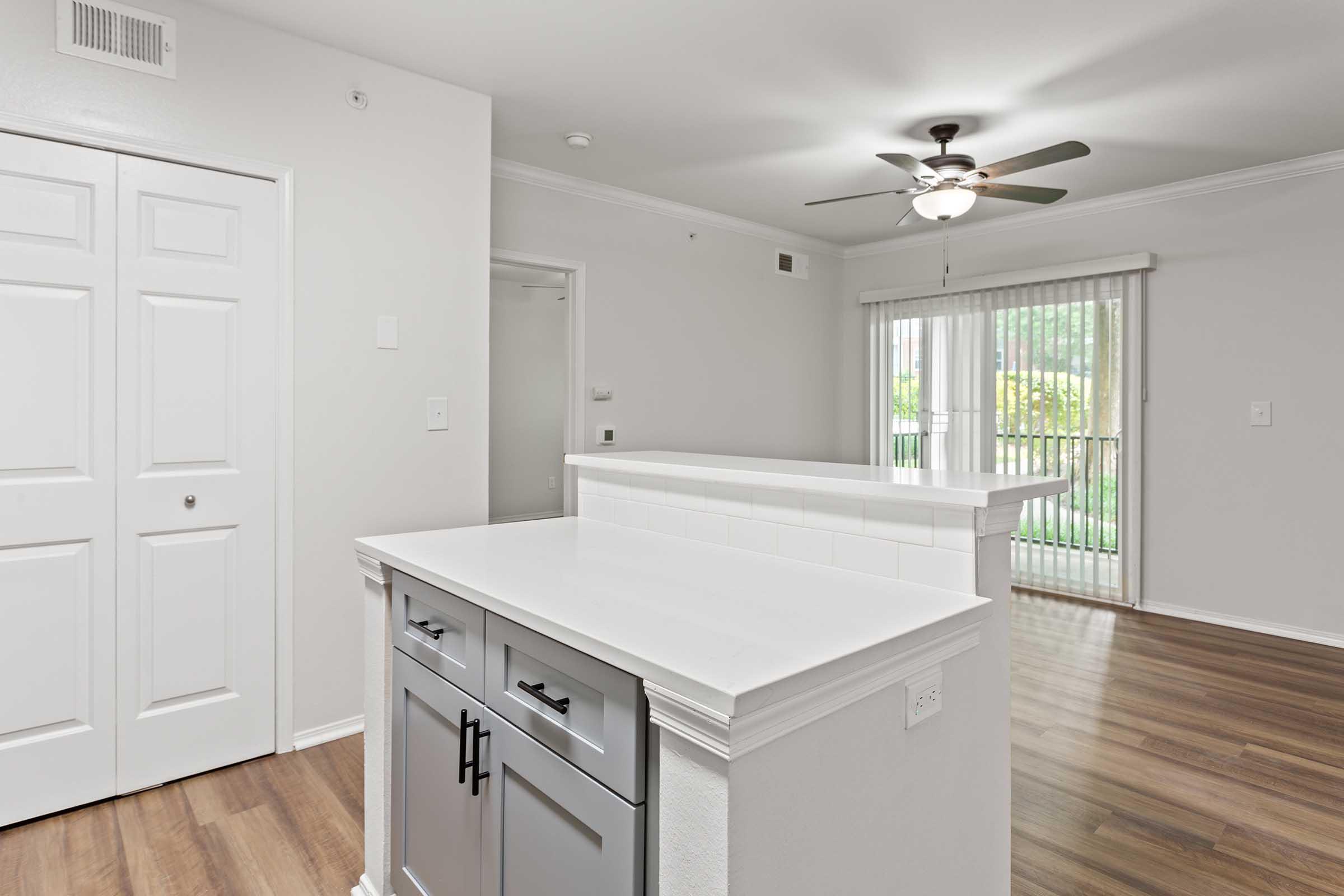 Interior view of a modern apartment featuring a kitchen with a white countertop and gray cabinetry. There is a closed door leading to another room, a ceiling fan overhead, and a sliding glass door with blinds visible in the background, leading to a bright outdoor area. The flooring is light-colored wood laminate.