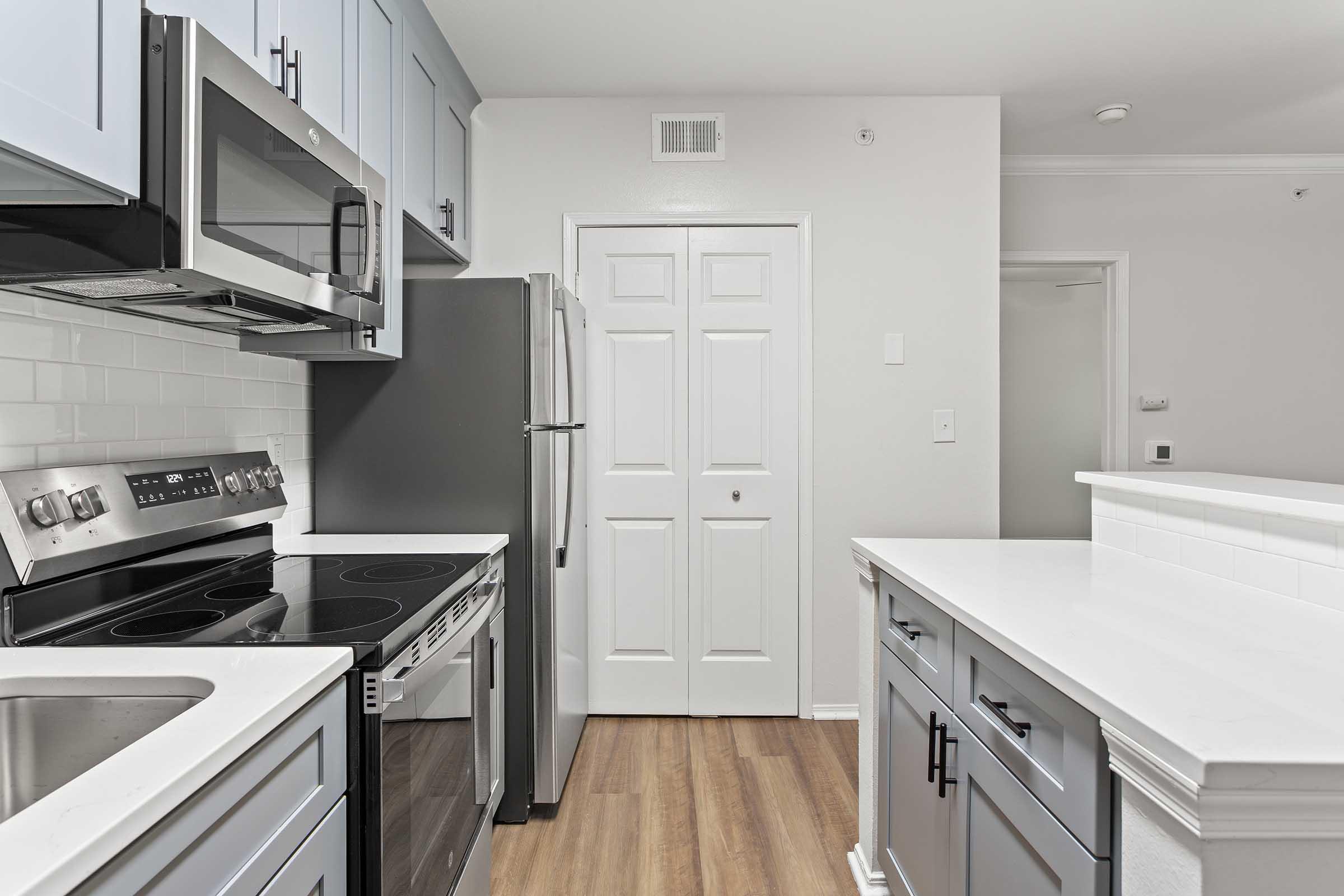 Modern kitchen featuring stainless steel appliances including a microwave, oven, and refrigerator. The cabinetry is a mix of light and dark tones, with a white countertop and a stylish backsplash. The space has wooden flooring and white walls, creating a bright and inviting atmosphere.