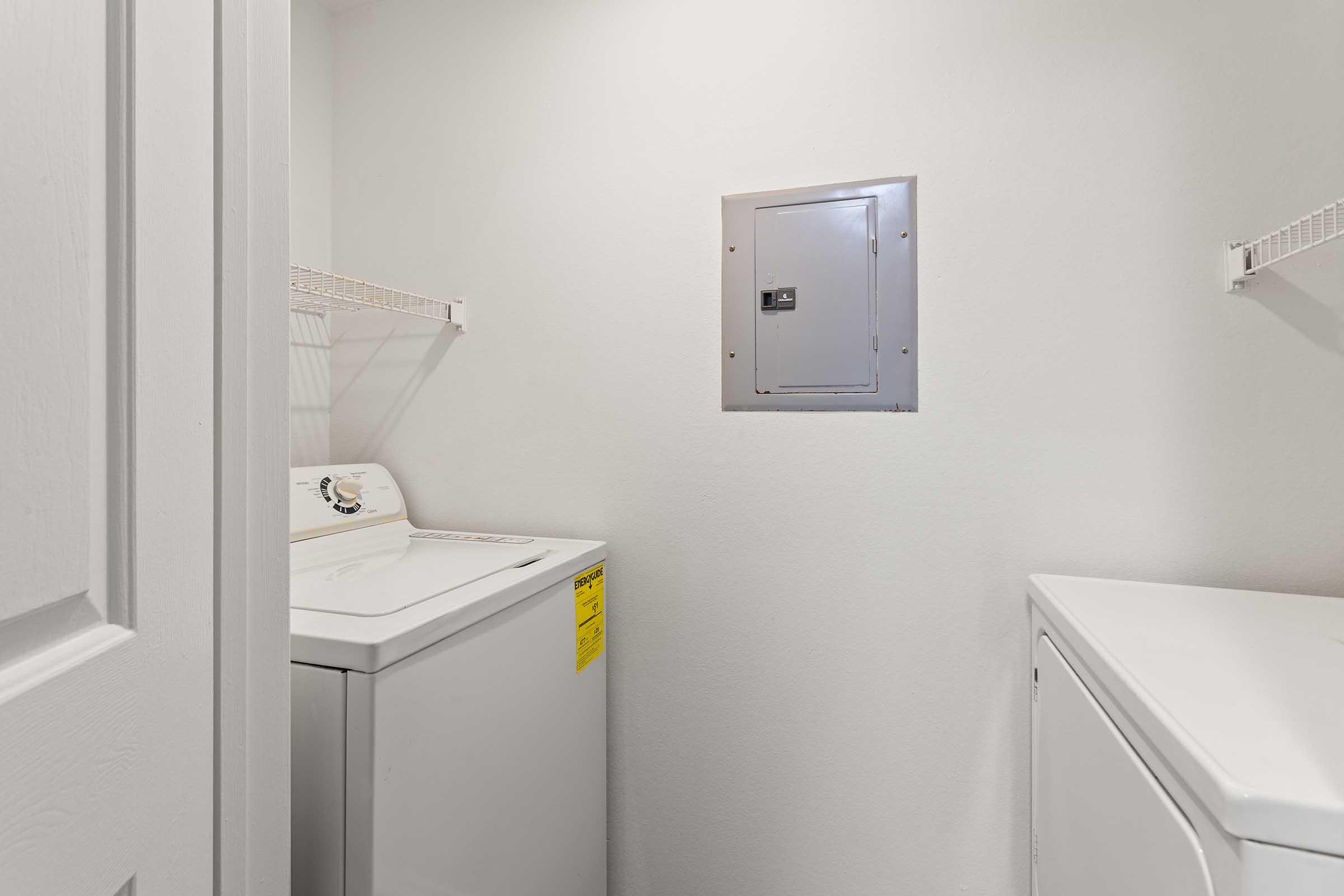 A laundry room featuring a washing machine and a dryer side by side. There is a utility panel on the wall and metal shelves above the appliances for storage. The walls are painted white, creating a clean and simple appearance.