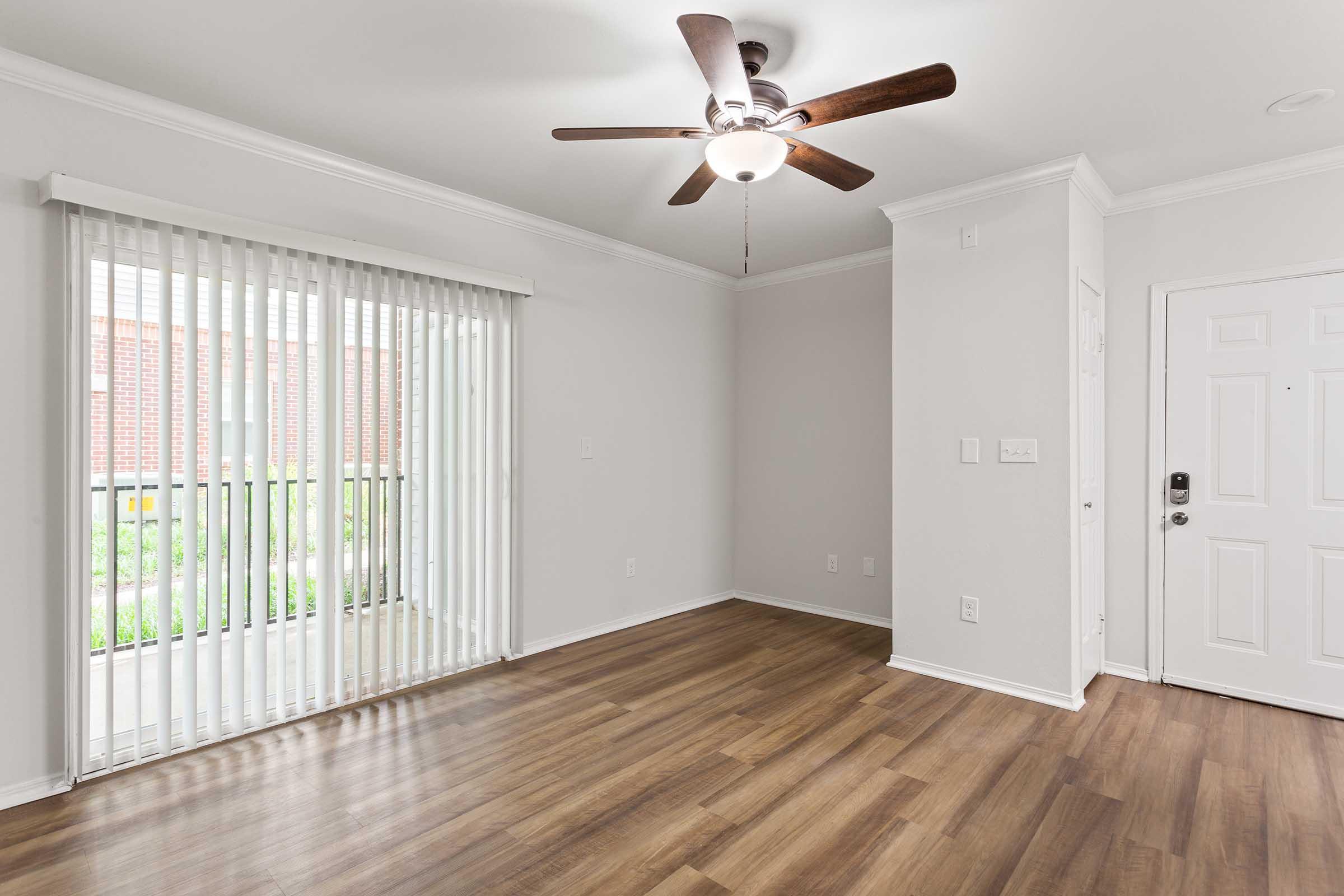 Bright and spacious room with light-colored walls and wooden flooring. A ceiling fan is mounted above, and large vertical blinds cover a window that looks out onto greenery. The entrance door is visible on the right, contributing to a welcoming atmosphere.