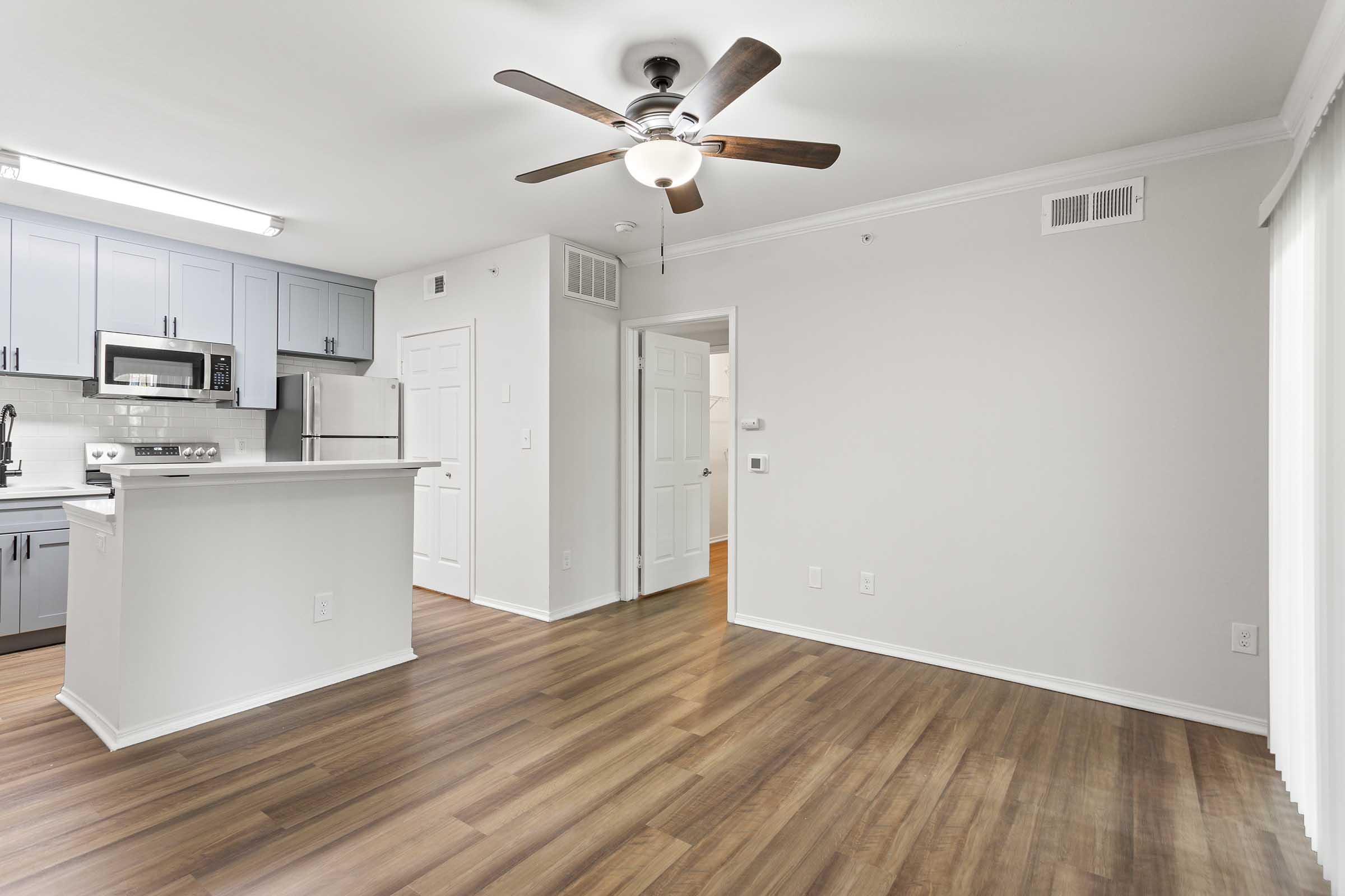 Modern kitchen and living area featuring wood flooring, a ceiling fan, and a light color palette. The kitchen includes gray cabinets, stainless steel appliances, and a countertop that divides the space. A door leads to another room, while large windows provide natural light.