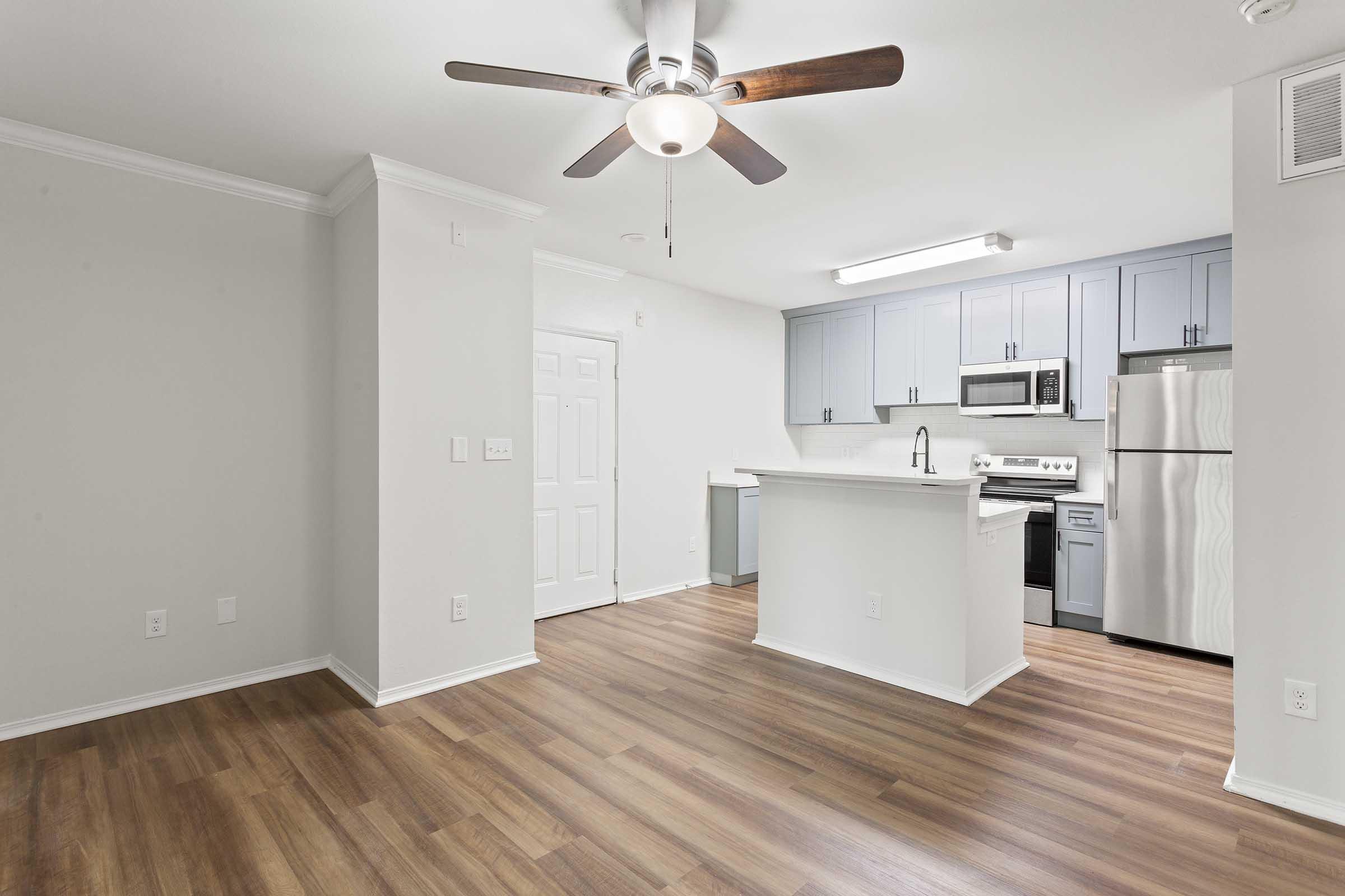 Modern kitchen and living area featuring light gray walls, a ceiling fan, and warm wood-like flooring. The kitchen has blue cabinets, stainless steel appliances, and a large island with seating. Natural light illuminates the space, creating a bright and inviting atmosphere.