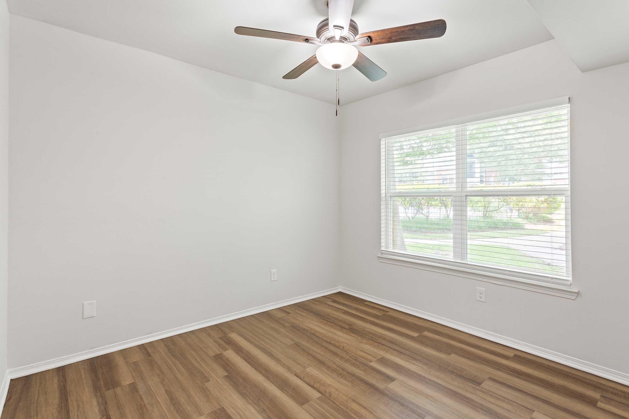 A spacious, empty room featuring light-colored walls and a wooden floor. A ceiling fan with wooden blades is installed above, and a large window with blinds allows natural light to illuminate the space. The room has a clean, minimalist design, suitable for various uses.