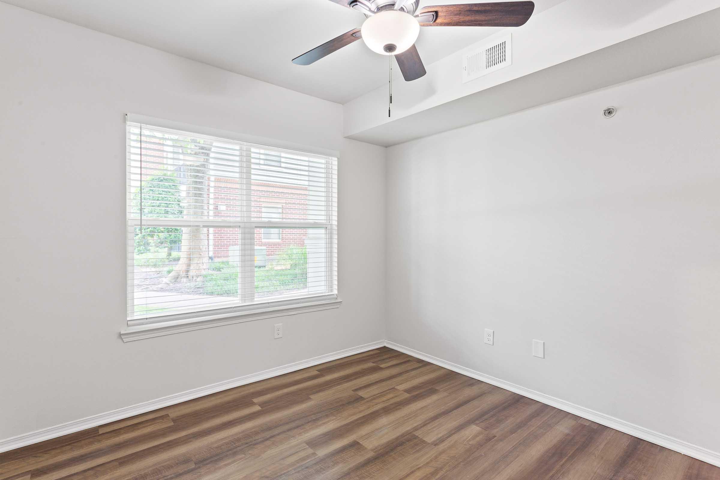 Bright, sparsely furnished room with light-colored walls and a ceiling fan. There's a window with blinds letting in natural light, revealing greenery outside. The flooring is wooden, and the space is ready for additional furniture or decoration.