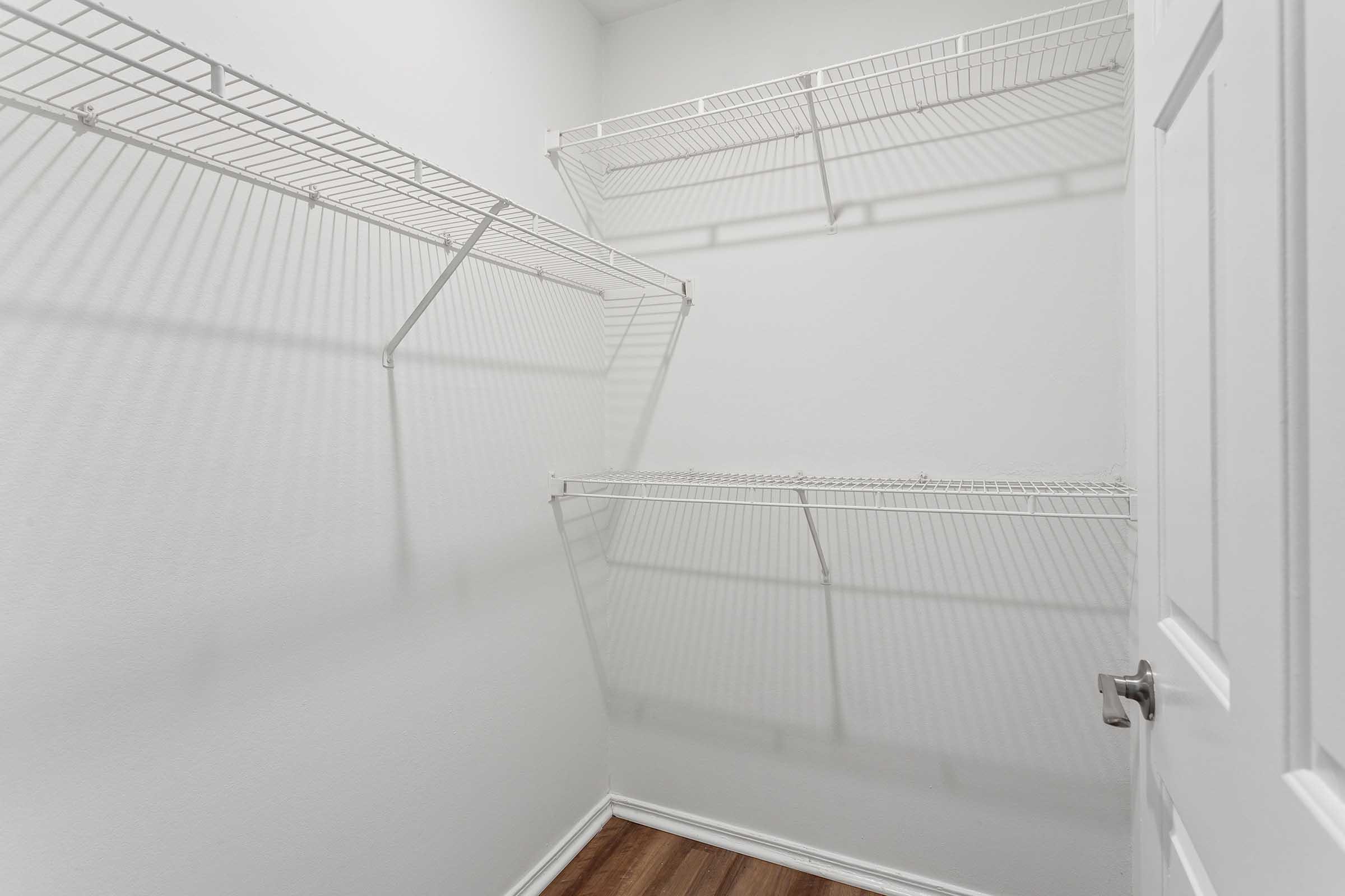 Empty closet with white wire shelving on two walls, light-colored walls, and hardwood flooring. The door is partially visible on the right side. Shadows from the shelves create a pattern on the wall.