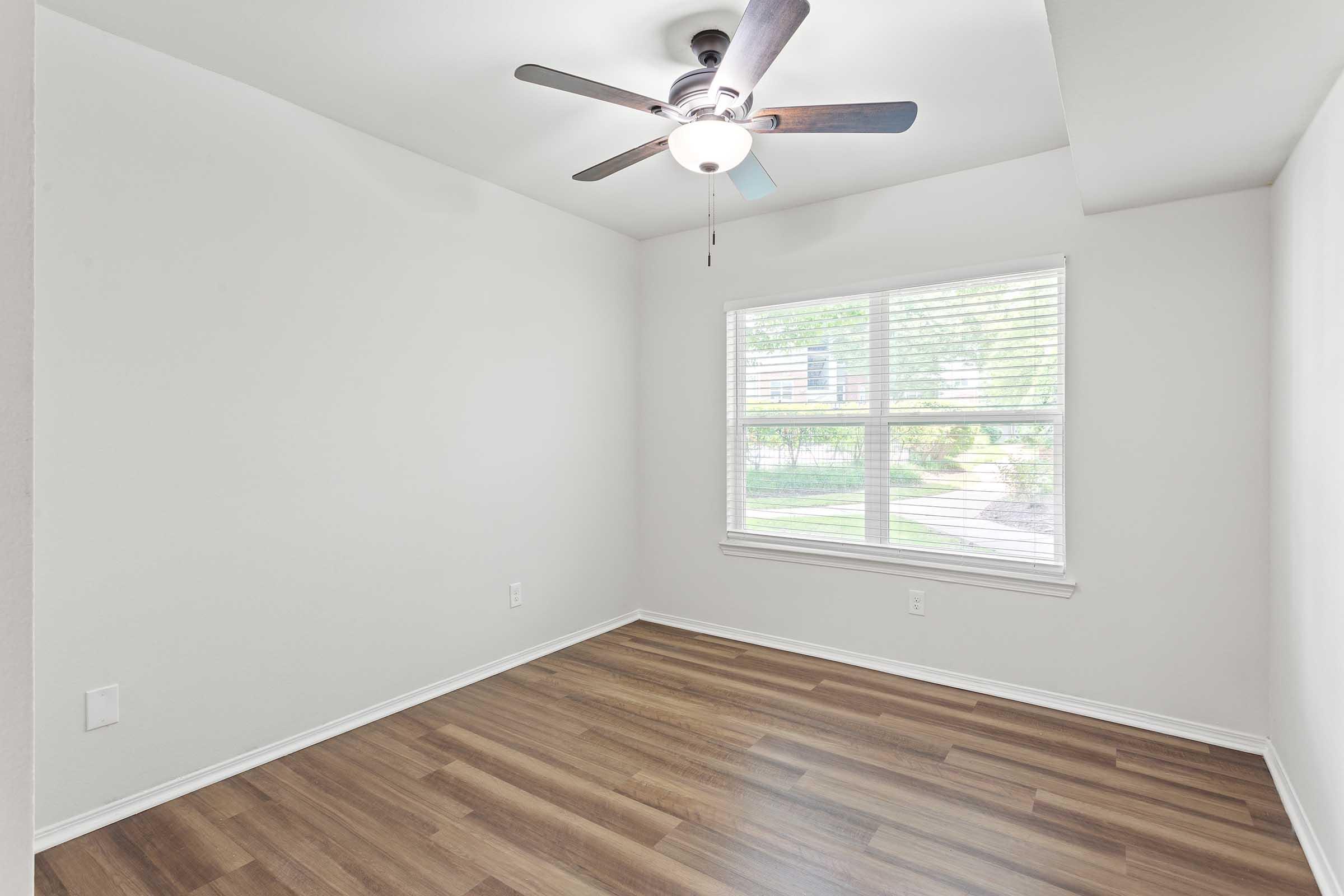 Empty room with light-colored walls and a ceiling fan. There is a window with blinds allowing natural light to enter, revealing a green outdoor view. The flooring is a wood laminate, and the space appears clean and well-maintained, suitable for various uses, like a bedroom or office.