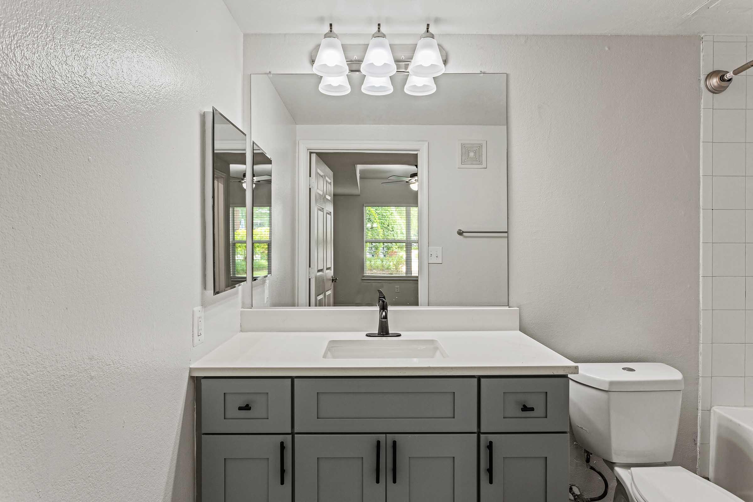 A modern bathroom featuring a gray vanity with a white sink, a large mirror above, and three pendant lights. The space includes a toilet and a tiled shower area, with a window allowing natural light to enter, highlighting a clean and contemporary design.