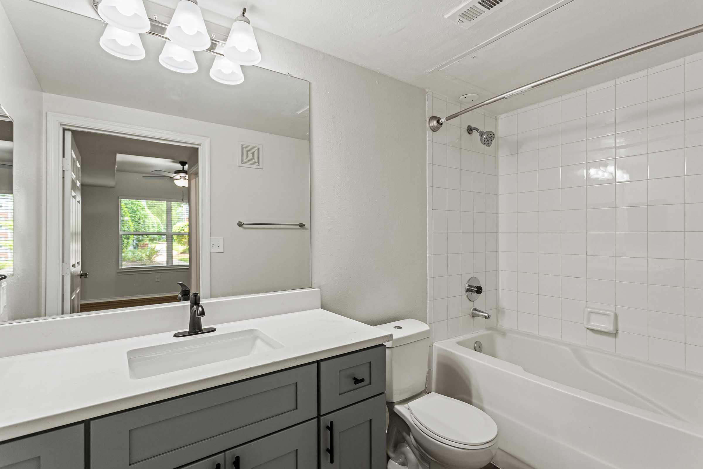Modern bathroom featuring a white tiled wall, a bathtub with a shower, and a sleek gray vanity. The sink area has a black faucet, and overhead lights illuminate the space. A door leads to a room with a ceiling fan and window, adding natural light to the overall design.