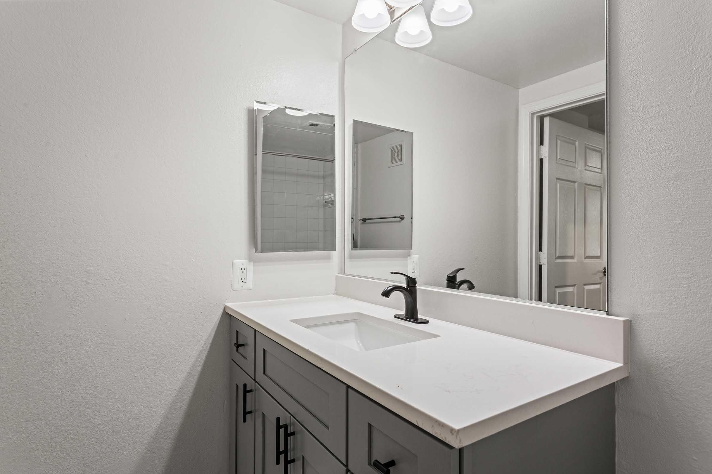 A modern bathroom featuring a white sink on a sleek gray countertop. The wall has a large rectangular mirror above the sink, with three light fixtures above it. A white door is partially visible in the background, and the walls are painted in a light gray color, creating a clean and contemporary atmosphere.