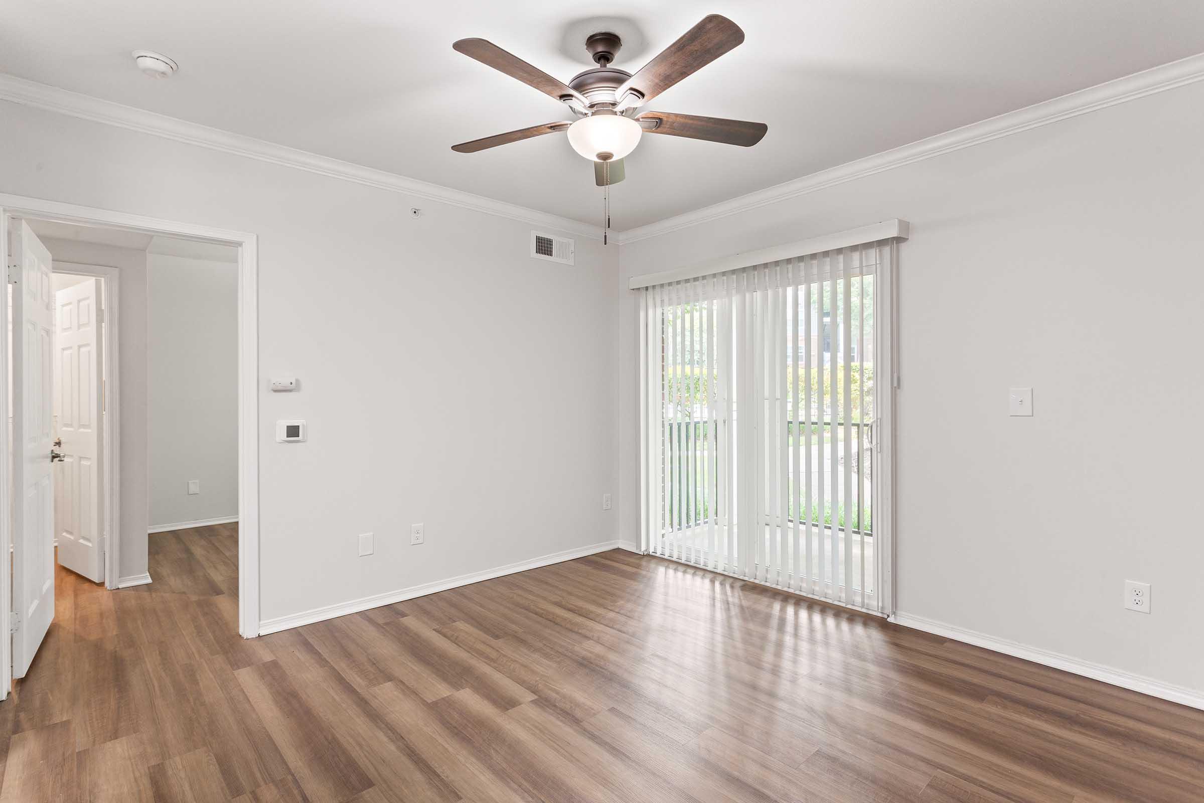 A well-lit room with light gray walls, hardwood flooring, and a ceiling fan. A sliding glass door with vertical blinds allows natural light. An entrance leads to another room, while a wall features a small control panel. The space appears clean and minimally decorated.