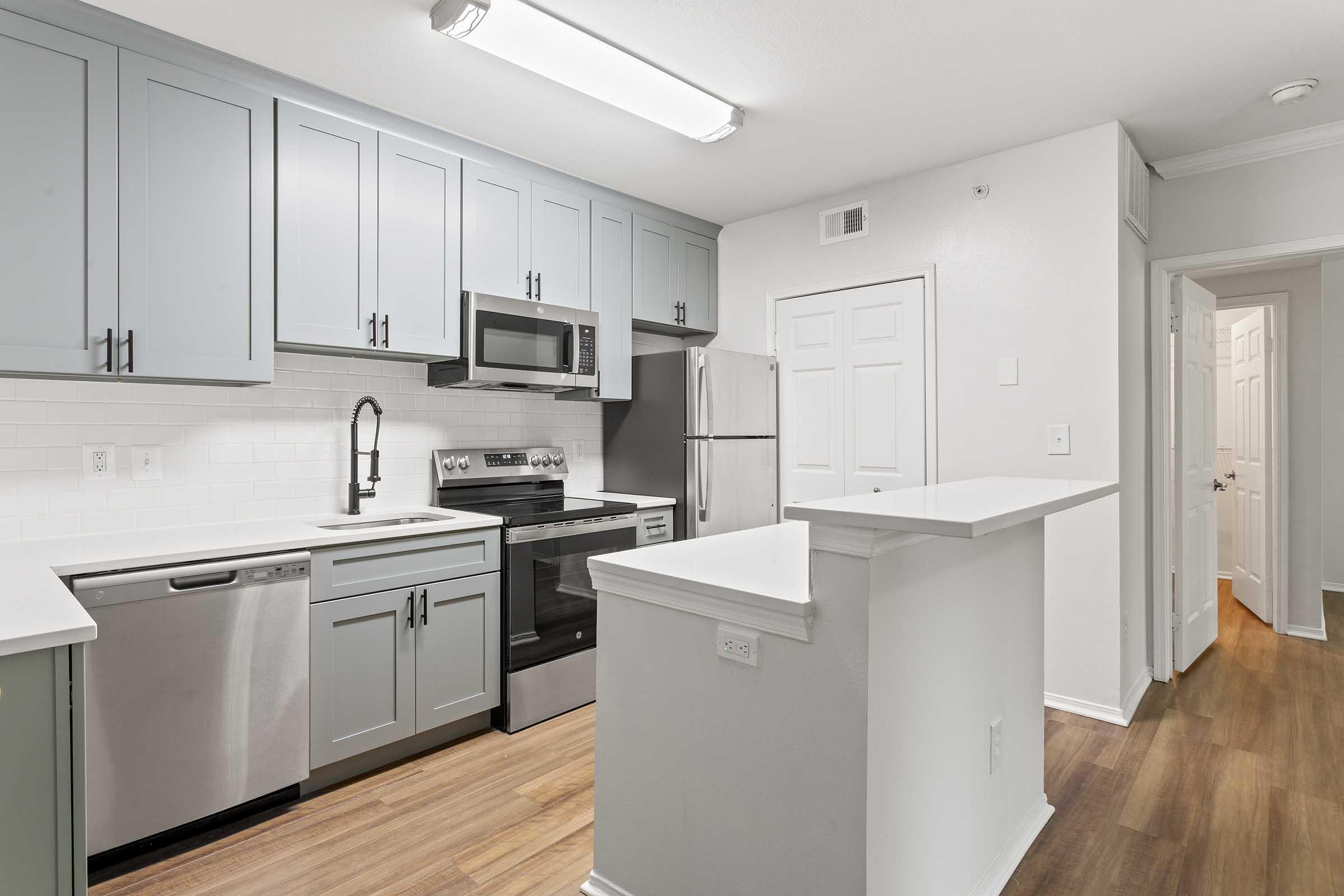Modern kitchen featuring light gray cabinetry, stainless steel appliances, and a white tile backsplash. Includes a sink, dishwasher, microwave, and a large countertop that separates the kitchen from an adjoining hallway. The space has wood flooring and bright, overhead lighting.
