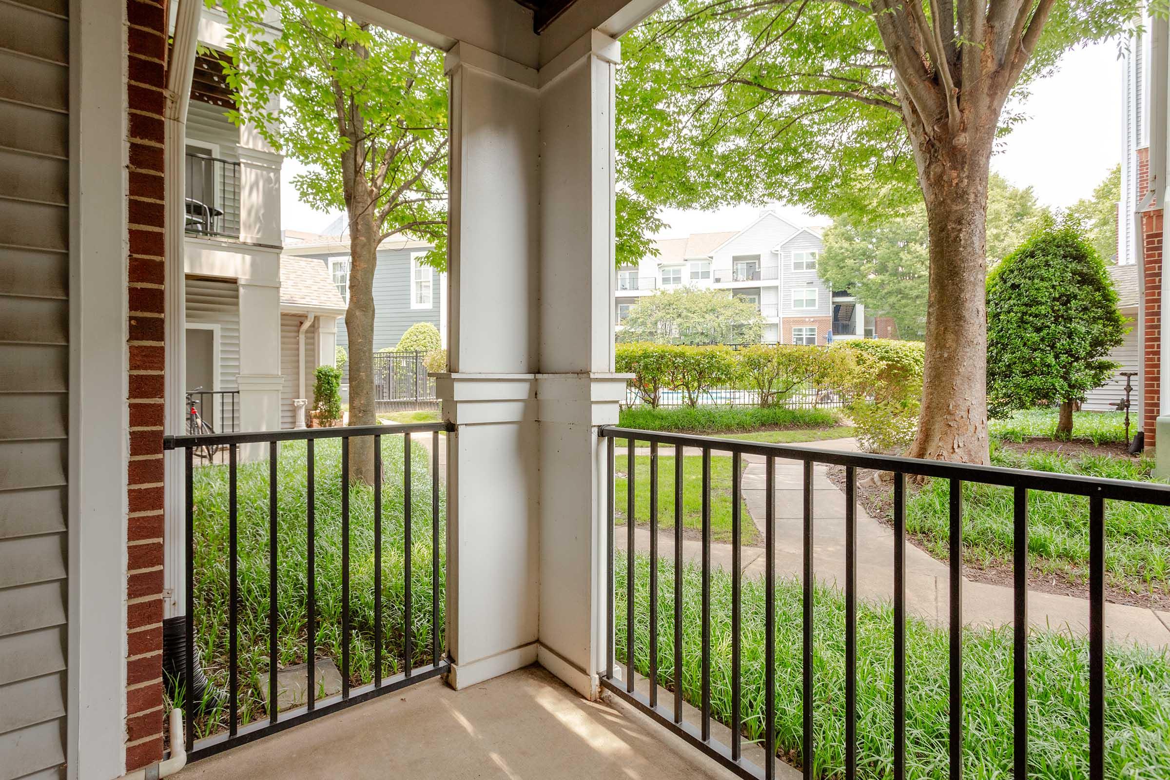 View from a covered porch showcasing a green courtyard with manicured bushes and trees, surrounded by apartment buildings. The scene conveys a calm and welcoming atmosphere, ideal for relaxation and enjoying the outdoors. The porch features black railings and light-colored walls.
