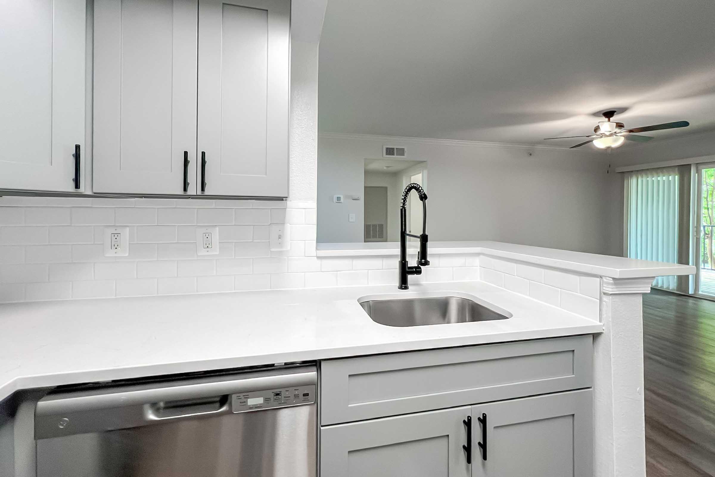 Modern kitchen featuring gray cabinets, a white countertop, a stainless steel dishwasher, and a sleek black faucet over a stainless steel sink. The background shows a neutral-colored living area with a ceiling fan and sliding glass doors, providing a bright, airy atmosphere.