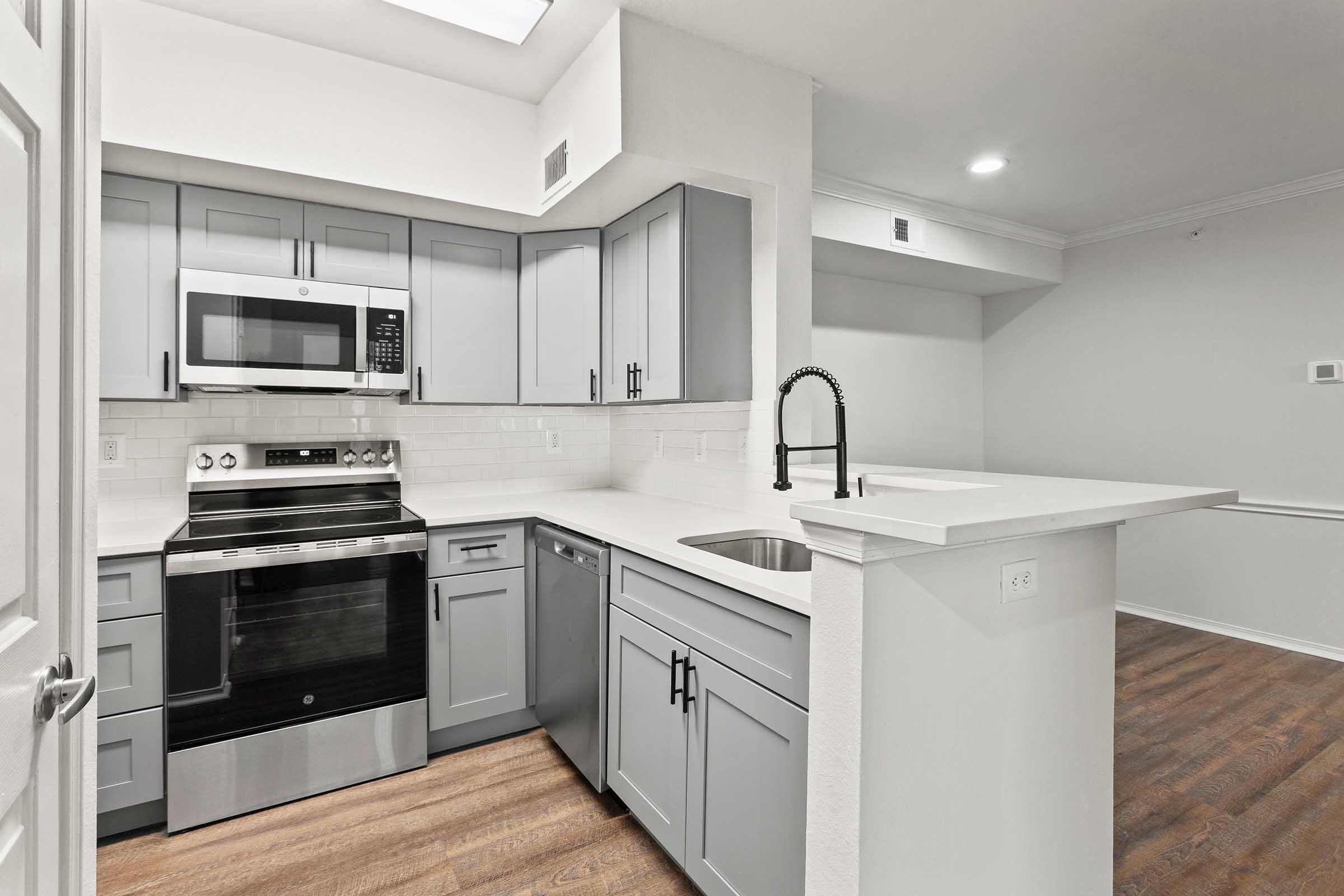 Modern kitchen featuring gray cabinets, stainless steel appliances, a microwave, and a countertop with a sink. The space is well-lit with overhead lighting, and there is an open layout connecting to a living area with hardwood flooring.