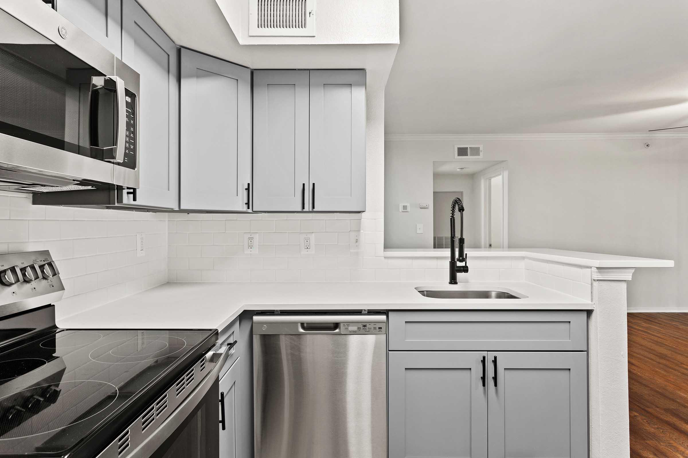 Modern kitchen featuring light gray cabinetry, a stainless steel microwave, oven, and dishwasher, with a sleek white countertop and a black faucet. The open layout connects to a brightly lit living area with hardwood flooring and neutral-colored walls.