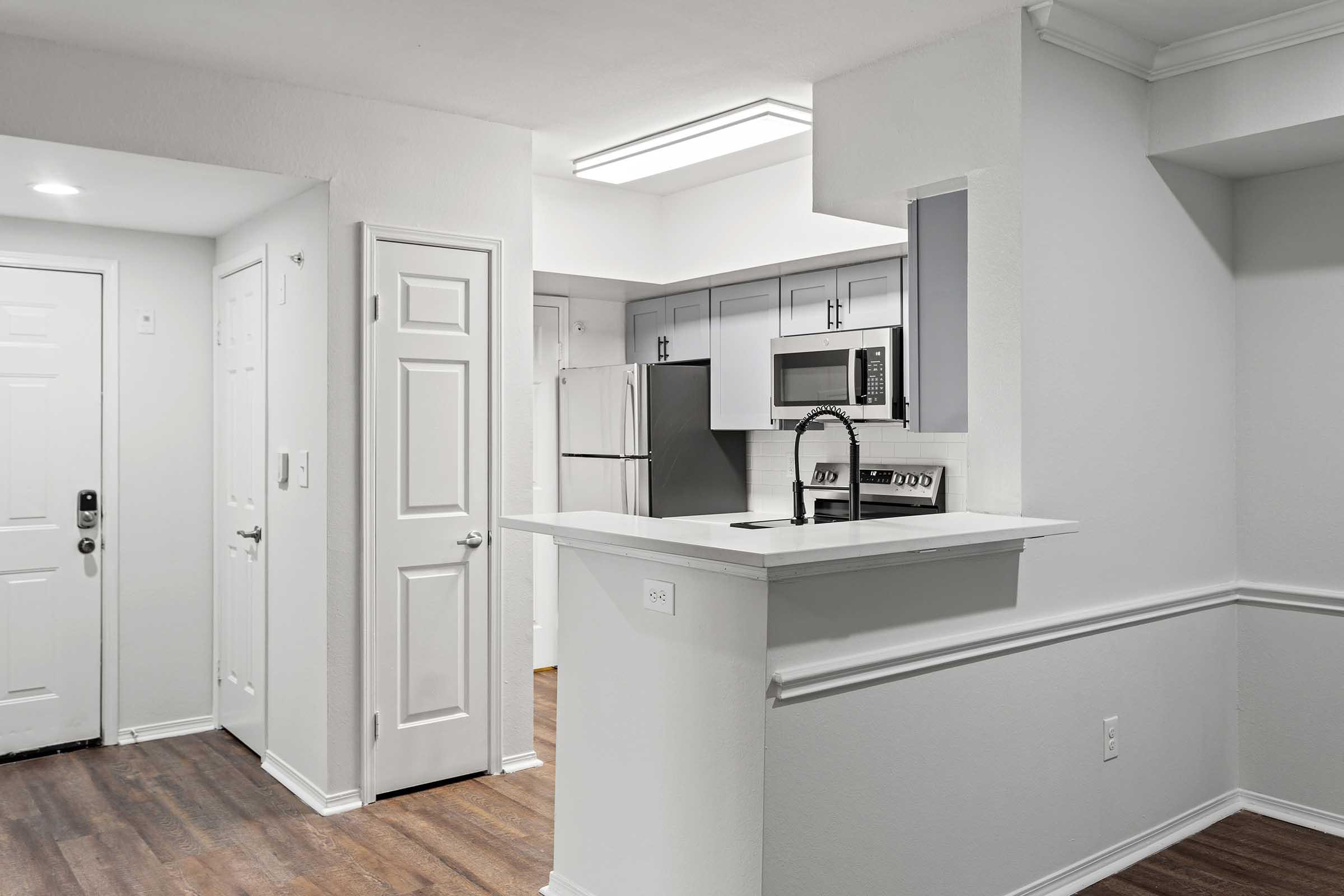 Modern kitchen and living area, featuring an open layout with white walls, a small breakfast bar, stainless steel appliances including a refrigerator and microwave, and wood-style flooring. The entrance door is visible on the left side, and there is ample natural light from the ceiling fixtures.