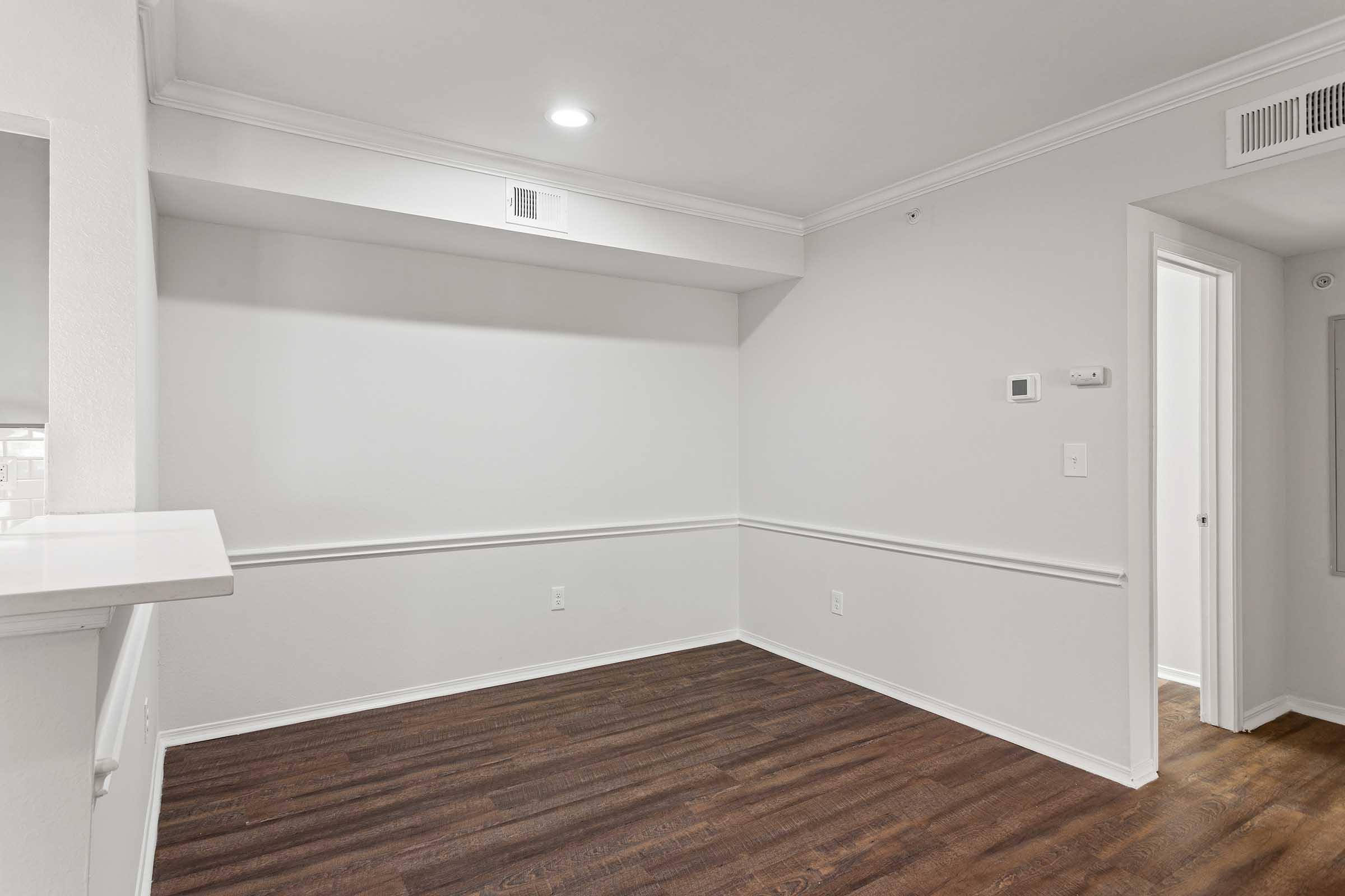 Empty room featuring light-colored walls, a wood-style floor, and crown molding. A small area with a wall-mounted thermostat is visible. There is a doorway leading to another room, and a counter space on the left side. Natural light comes from an overhead light fixture.