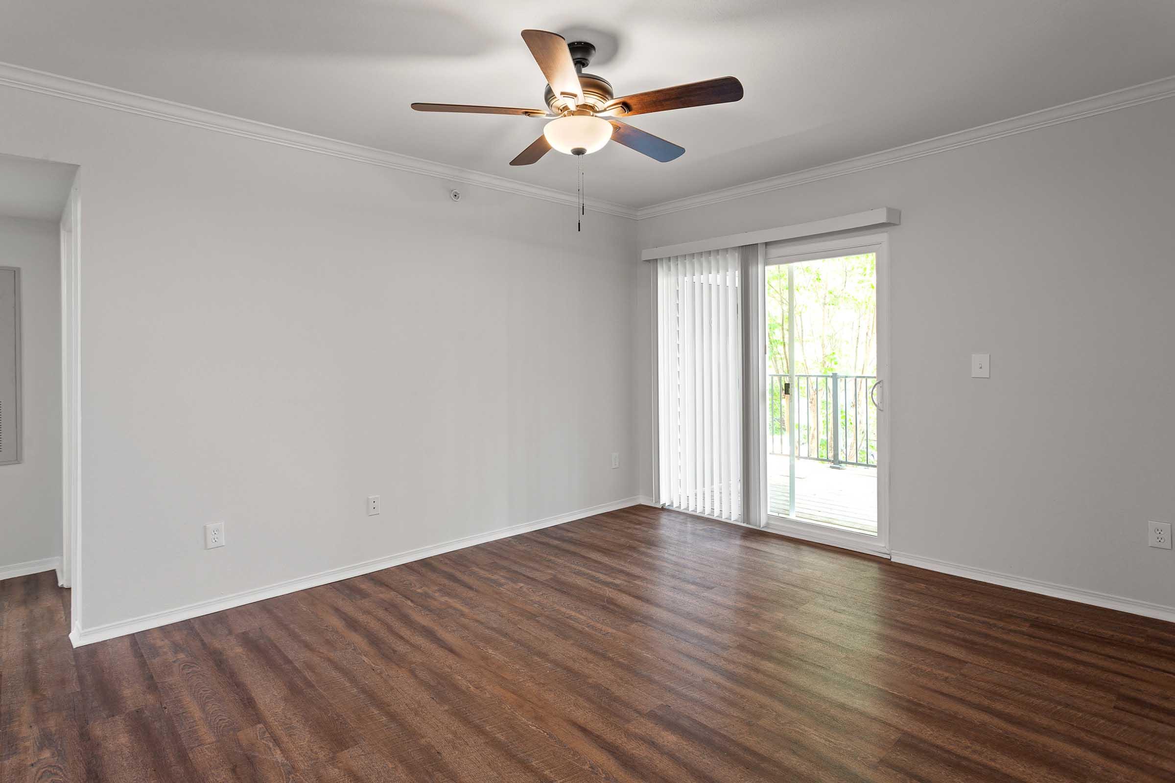 A spacious, empty living room with light gray walls and hardwood flooring. It features a ceiling fan and large sliding glass doors that lead to a balcony. Natural light floods the room, highlighting the simplicity and openness of the space.