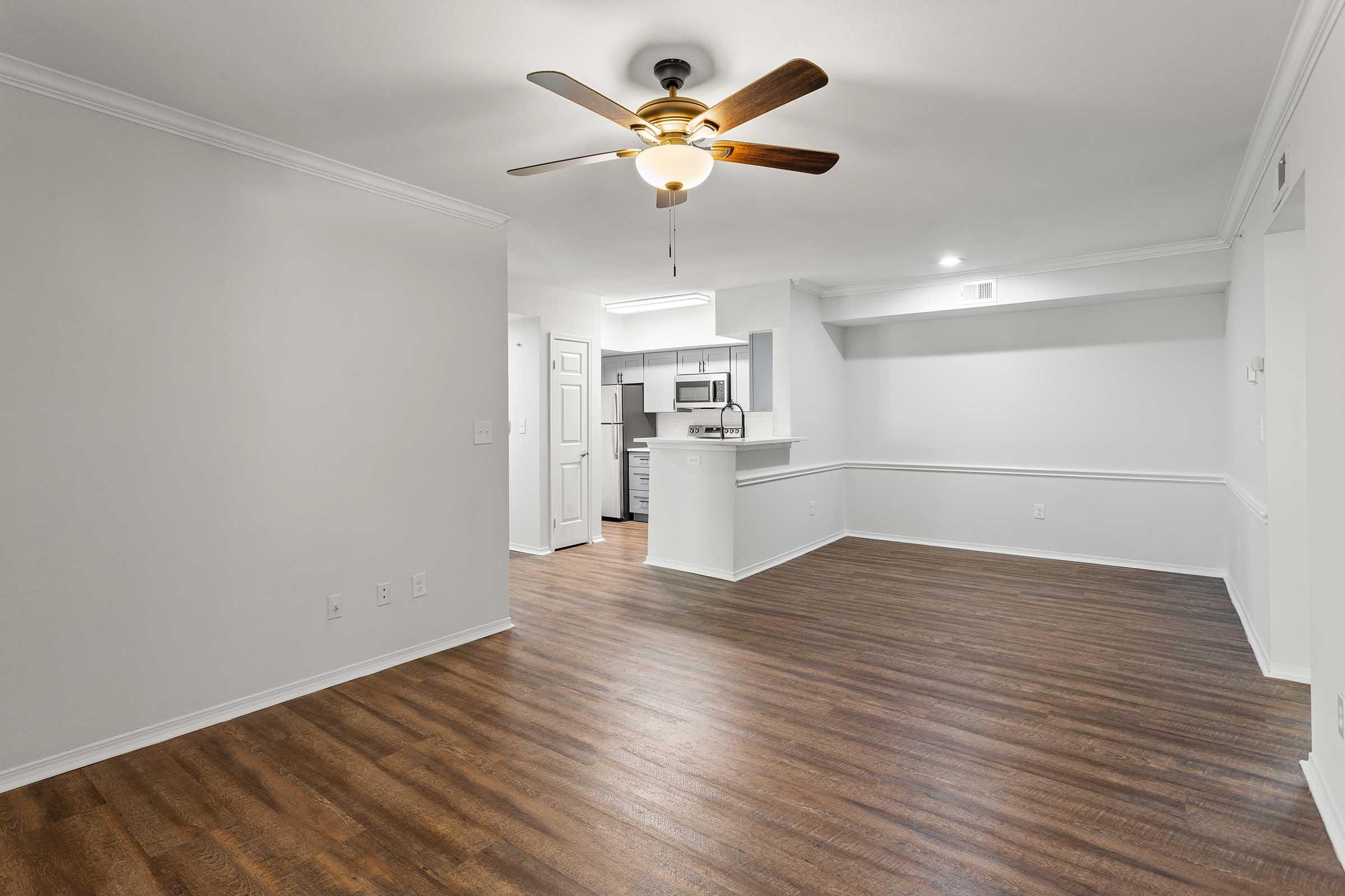 A spacious, light-colored living area with a ceiling fan and hardwood flooring. To the left, there is a small kitchen area visible with modern appliances. White walls create a bright atmosphere, and a half wall separates the living space from the kitchen. Natural light illuminates the room.