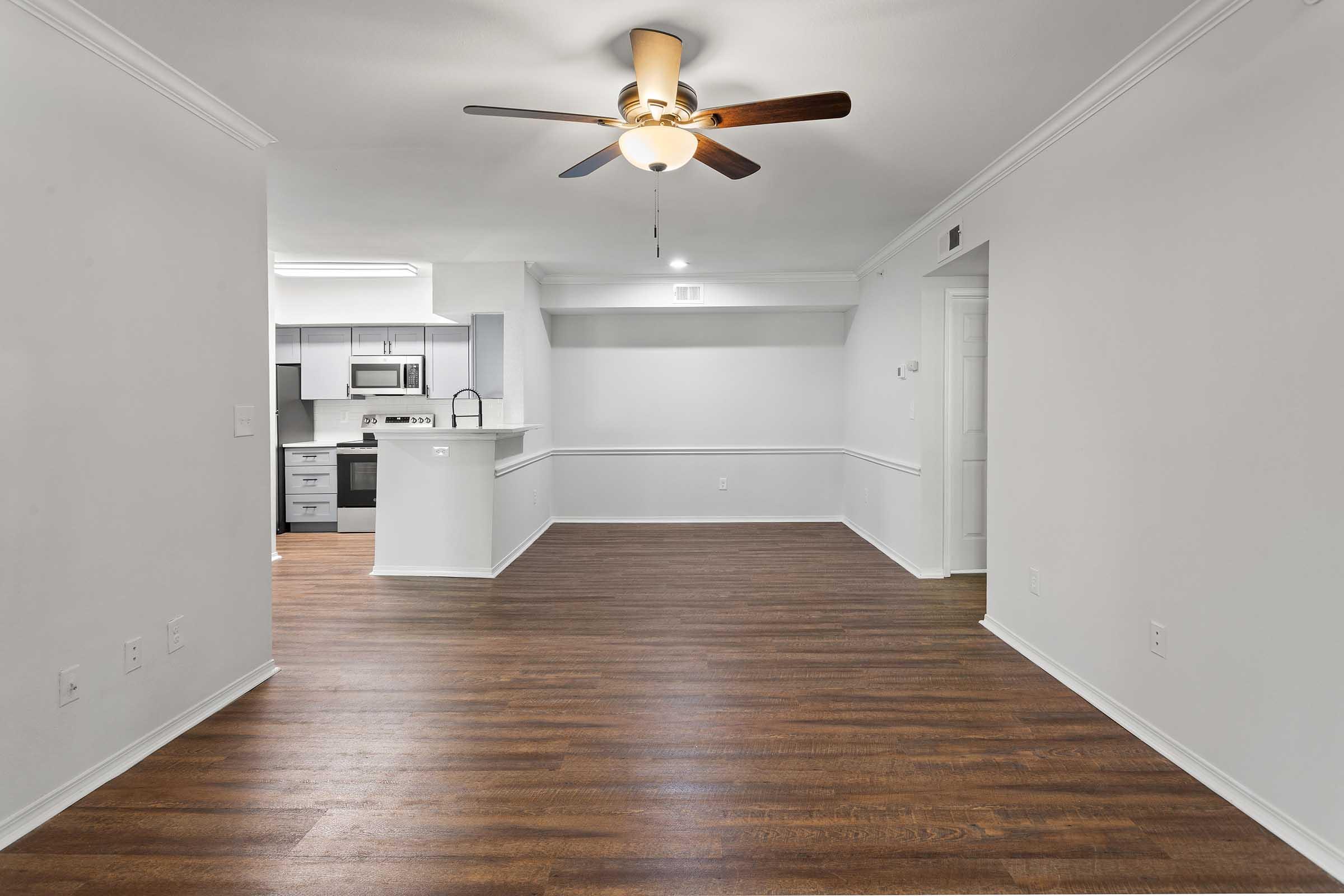 A spacious and modern living area featuring a ceiling fan, light-colored walls, and hardwood flooring. On one side, there is an open kitchen area with stainless steel appliances and a breakfast bar. The layout offers a bright and inviting atmosphere, suitable for various furniture arrangements.