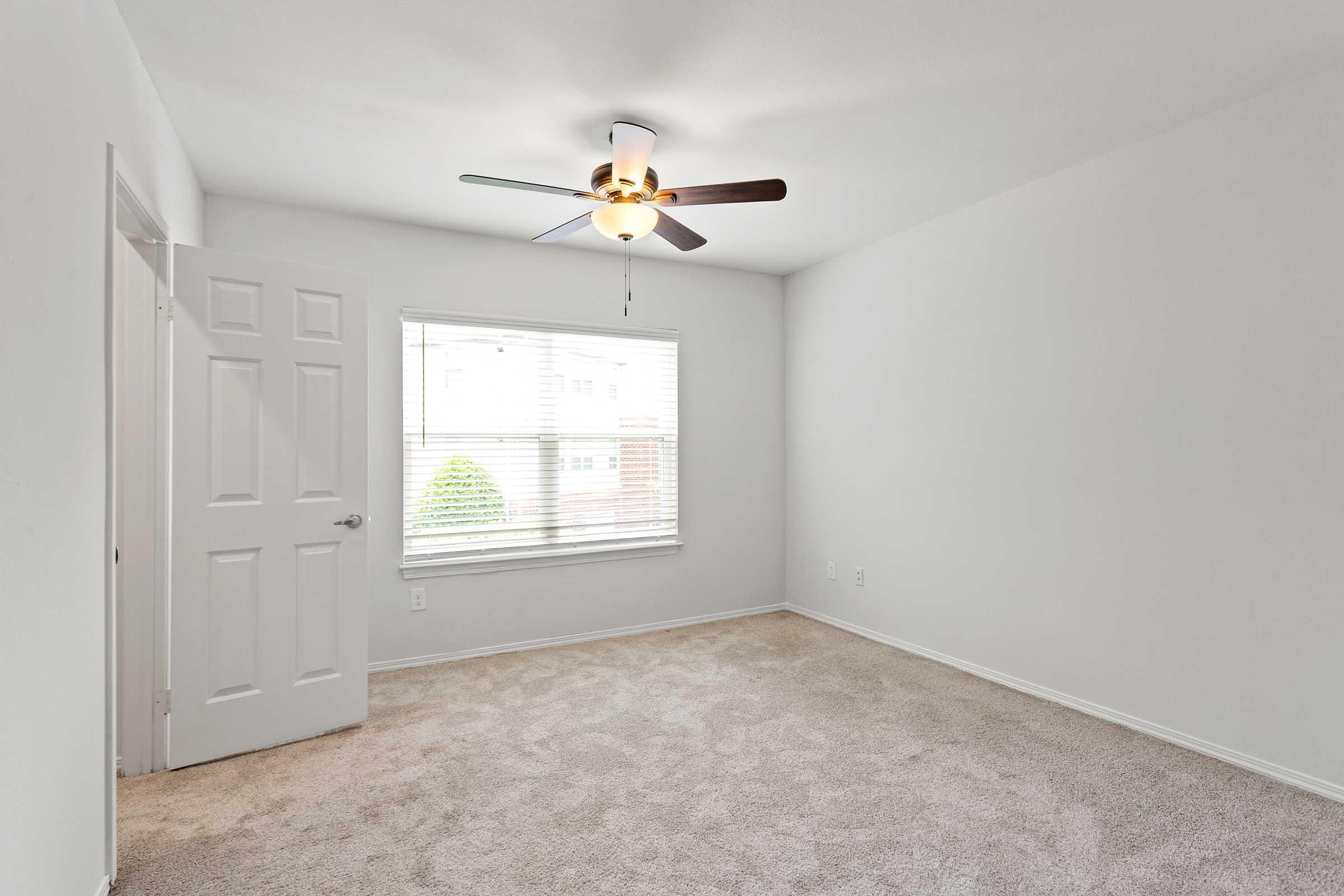 A light and airy bedroom with beige carpet, a ceiling fan, and a large window allowing natural light. The walls are painted in a neutral tone, and there is a doorway leading to another room. The space appears clean and minimalistic, suitable for various decor styles.
